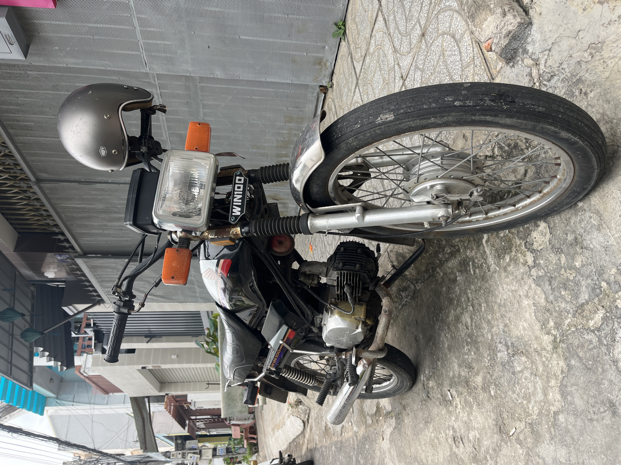 In Phu Thuy Ward, Vietnam, a black Honda Win motorcycle is parked on a concrete sidewalk. The motorcycle is facing the viewer, with its front wheel slightly turned to the left. A silver helmet rests on the handlebars. The motorcycle is equipped with an orange turn signal on the right side, a chrome exhaust pipe, and a visible engine. The surrounding environment appears to be an urban street with buildings and a metal fence visible in the background. The sidewalk is made of patterned concrete tiles. There are two cigarette butts on the ground near the motorcycle.