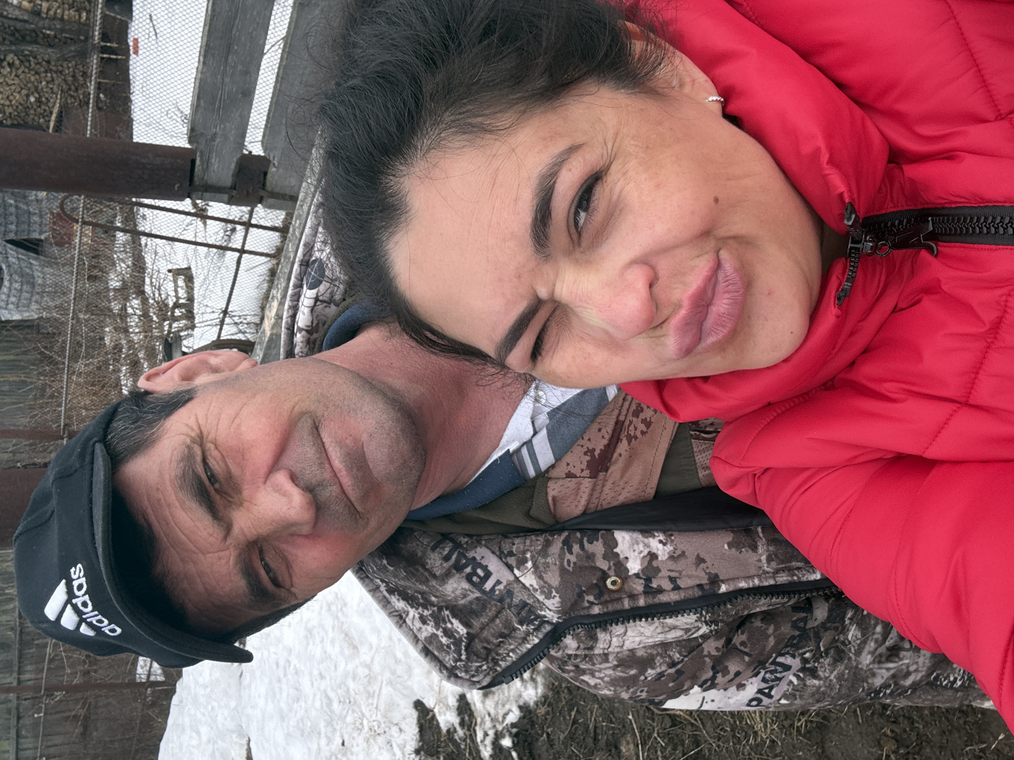 Two people, a man and a woman, are close together in an outdoor setting. The woman, on the right, has dark hair and is wearing a red puffer jacket. She is making a playful expression, squinting one eye and puckering her lips. The man, on the left, is wearing a black Adidas baseball cap and a camouflage-patterned jacket. There is some snow visible on the ground. The location is in Кемуль, Russia.