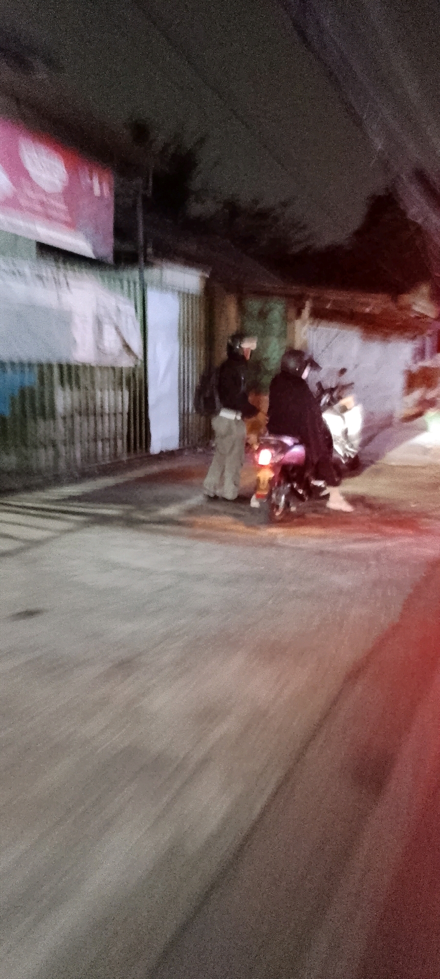 In Karangsatria, Indonesia, at night, two individuals on a motorbike are paused on a street, near a fence and building. The person in the foreground, dressed in light-colored pants and a dark jacket, stands next to the motorbike. The second person, wearing a dark outfit, is seated on the motorbike. The scene is characterized by low light conditions, with the motorbike's tail light emitting a red glow. The background features a fence with vertical metal bars and a portion of a building with a tarp. The street is paved and shows signs of wear.