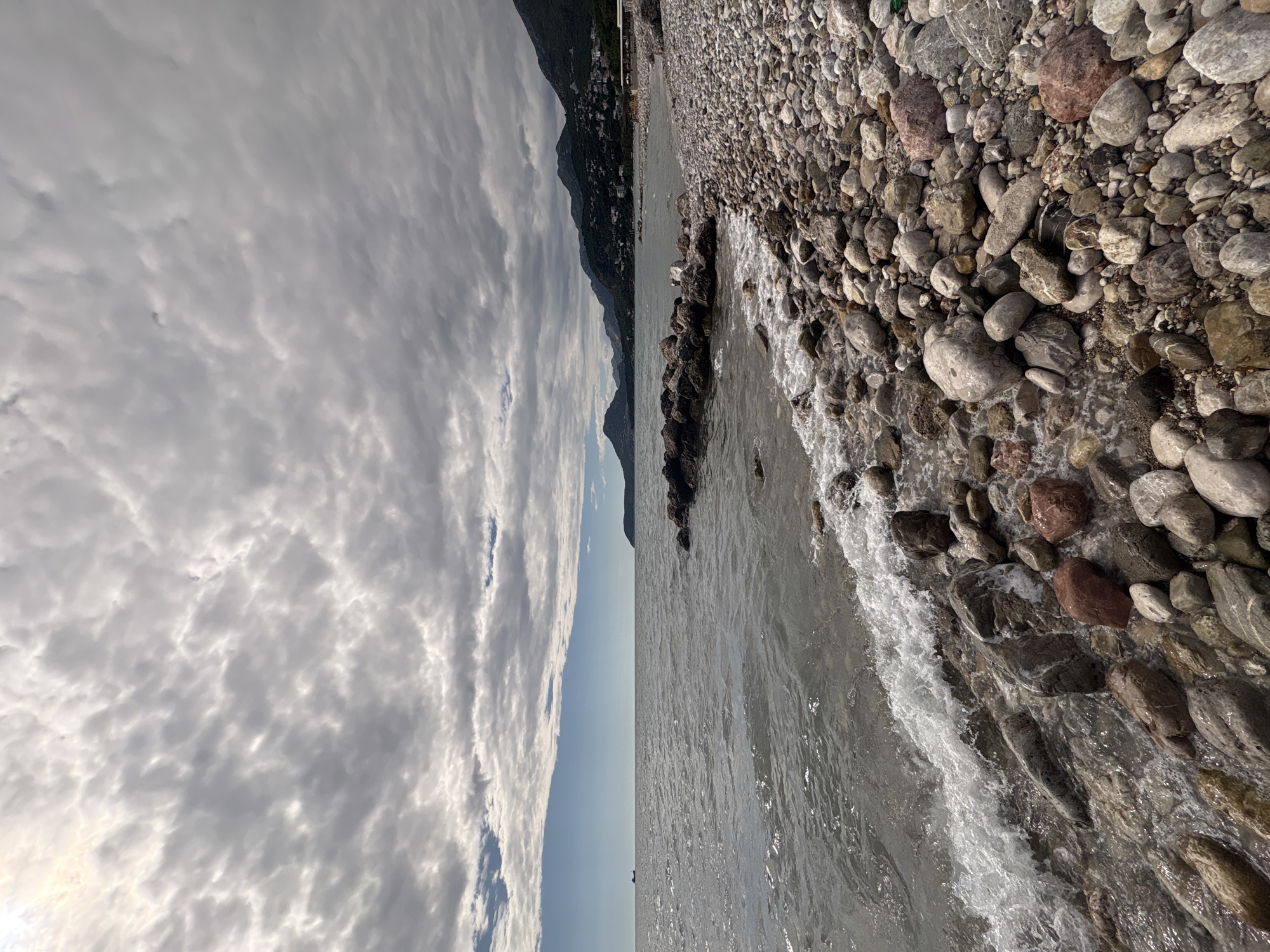 The image presents a panoramic coastal landscape in Bar, Montenegro, on an overcast day. In the foreground, a dense rocky beach is visible, covered with numerous pebbles and stones of various sizes, shapes, and colors, many appearing wet from the encroaching water. Gentle waves are breaking onto the shore, creating white foam along the waterline.
Extending into the midground from the beach is a line of larger, dark rocks, possibly a natural formation or a small breakwater, with the calm, rippling sea stretching beyond it. On the opposite side of the sea in the midground, towering mountains rise dramatically from the coastline. Hints of distant buildings or a small settlement can be discerned on the lower slopes of these mountains.
The sky, occupying a significant portion of the background, is dominated by a thick layer of textured, grey and white clouds, likely stratocumulus, which diffuses the daylight. The lighting suggests it is daytime. No people are visible in the scene, and there is no discernible text. The overall impression is one of rugged natural beauty and tranquility.