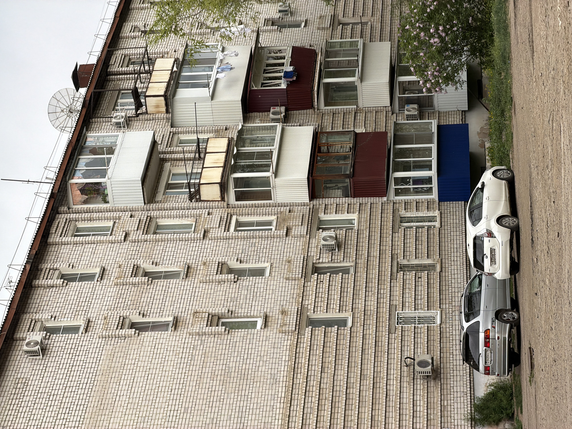 The image depicts a multi-story apartment building in Birobidzhan, Russia. The building is constructed of light-colored brick. Many of the apartments have enclosed balconies, some of which are painted white, maroon, or blue. Several satellite dishes are mounted on the roof. Air conditioning units are visible on the building's facade. Two cars are parked along the curb in front of the building. One is white and the other is silver. There are some trees on the right side of the image.