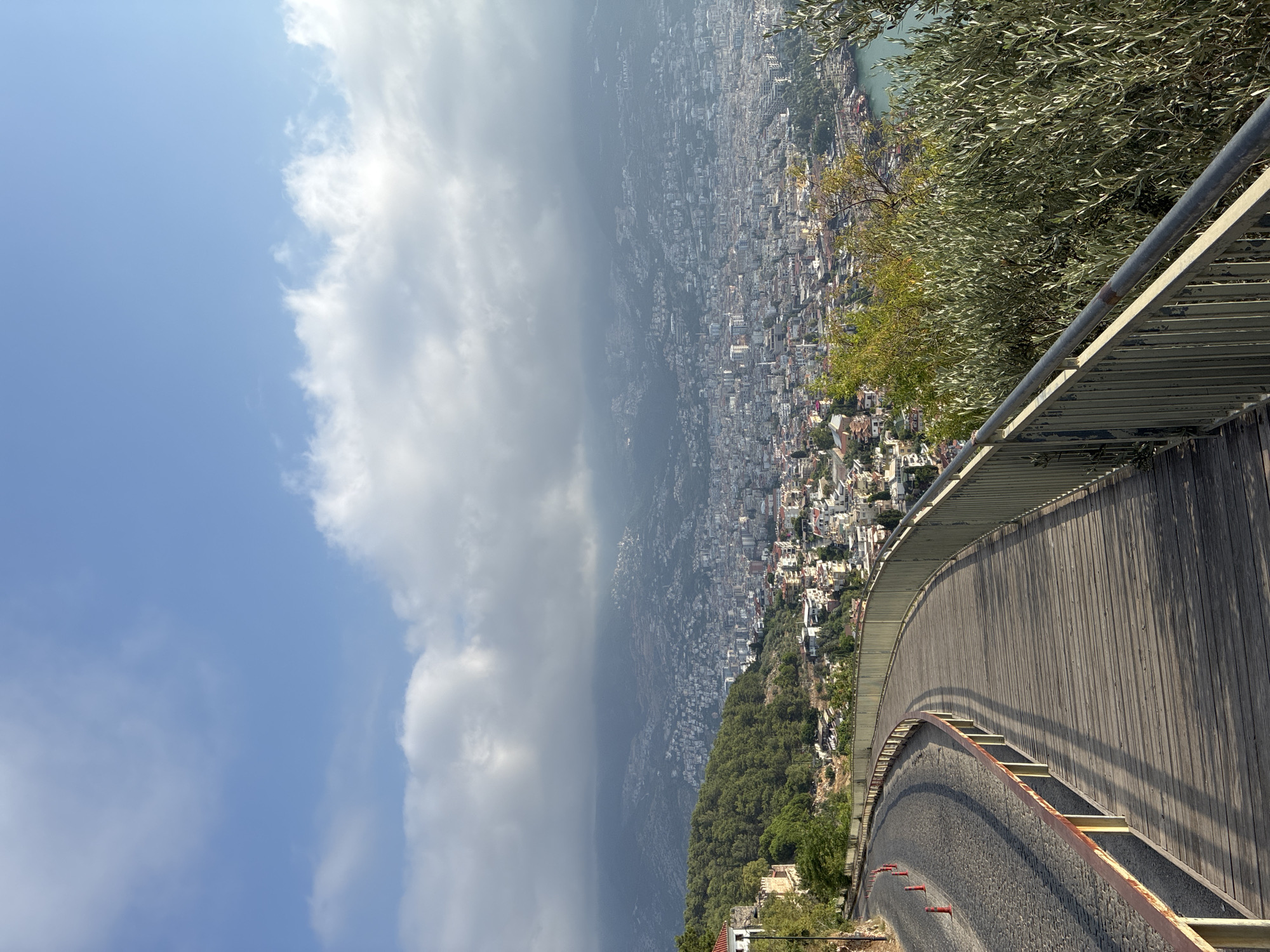 The image shows a scenic overlook of Alanya, Turkey. In the foreground, a curved wooden boardwalk with a metal railing runs along the edge of a steep incline. To the left of the boardwalk, a cobblestone path is visible, lined with orange traffic cones. Beyond the boardwalk and path, a sprawling city of white and terracotta buildings cascades down a hillside towards the sea. Lush green trees cover the lower slopes, and a hazy mountain range forms the backdrop. The sky is a vibrant blue, with scattered white clouds. The overall scene suggests a peaceful and picturesque view of the coastal city.