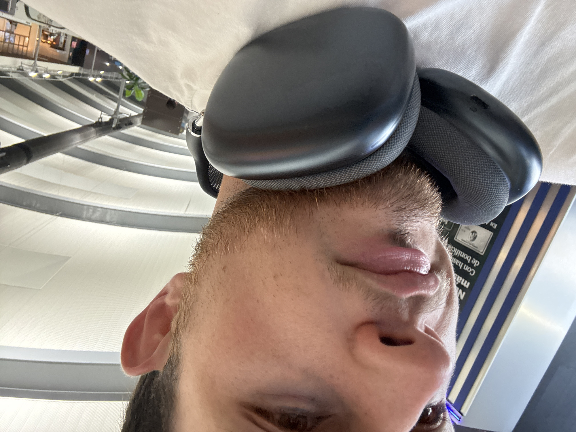 A close-up, low-angle shot captures the lower half of a man's face and head, with large black headphones positioned around his neck. The man has short, light brown stubble on his chin and jawline, and his lips are slightly parted. He is wearing a white t-shirt. In the background, a curved ceiling with white and gray stripes suggests an indoor public space, possibly an airport terminal. To the left, a tall, narrow object with blue and white vertical stripes is visible. The overall setting appears to be a modern, well-lit environment. The location is identified as Apodaca, Mexico.