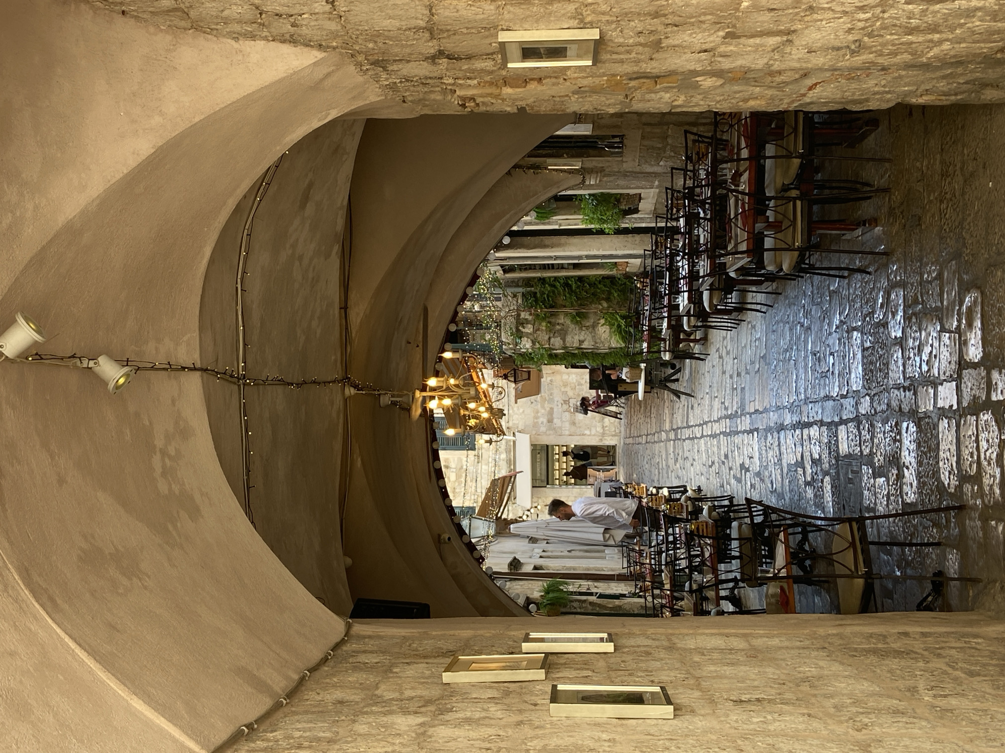 The image depicts an outdoor seating area of a restaurant in Dubrovnik, Croatia, likely within the Old Town. The scene is framed by an arched stone passageway, characteristic of Dubrovnik's ancient architecture. Rows of tables and chairs are set up along a cobblestone path, which appears wet, possibly from recent rain or cleaning. The lighting fixtures, including strings of small bulbs and an ornate chandelier, suggest that the establishment is prepared for evening service. Several framed pictures hang on the stone walls lining the passageway. A man in a white shirt stands near the tables, and other patrons are visible further down the walkway. Lush greenery is visible on some of the buildings in the background.