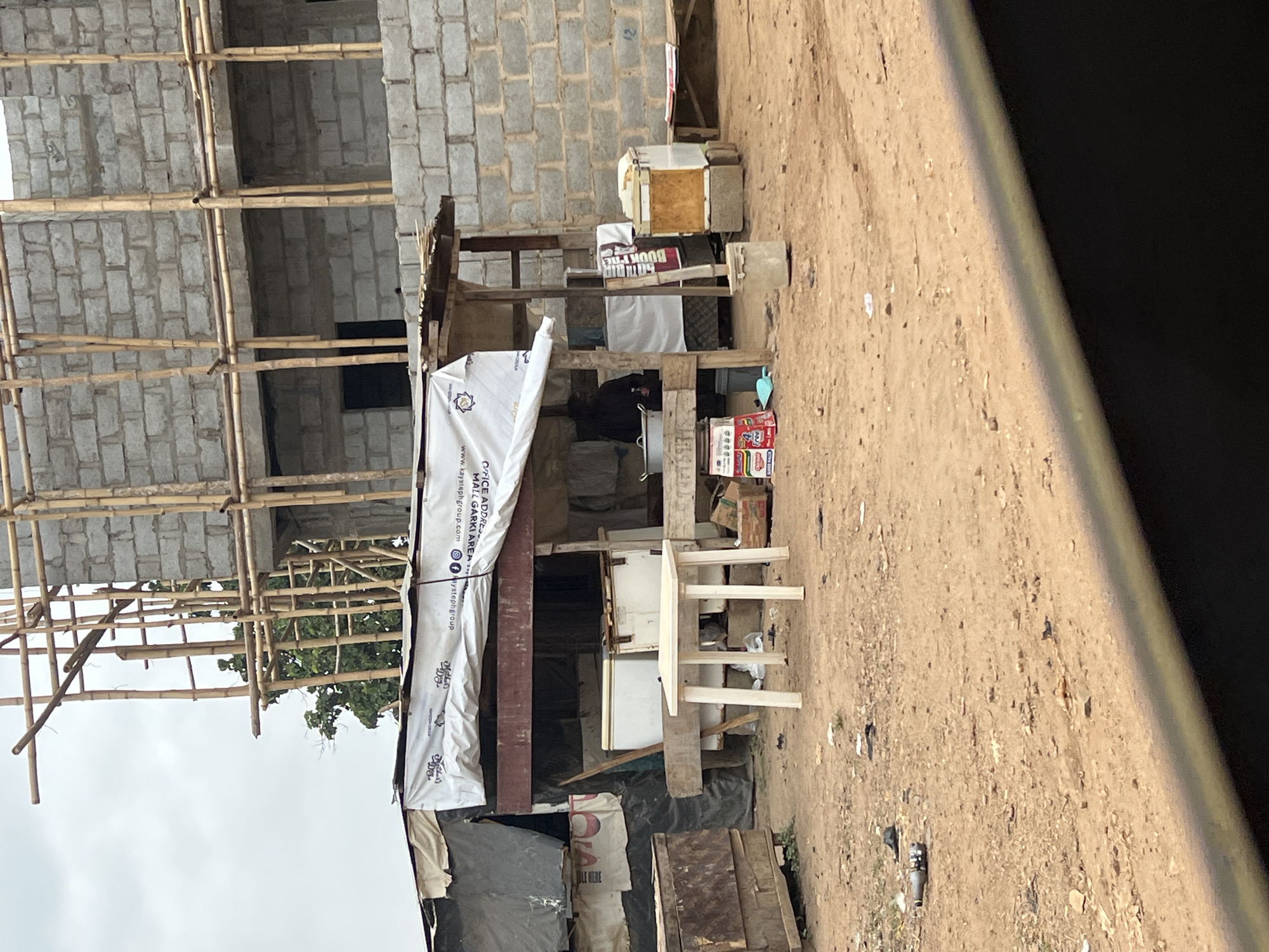 The image depicts an informal outdoor scene in what appears to be a developing area, likely Abuja, Nigeria as per the context. In the mid-ground, a makeshift stall or shop is set up under a simple wooden frame structure, covered partly by tarpaulin and banners. Inside the stall, a person dressed in dark clothing is partially visible, seated behind a counter. Various items commonly found in a small shop are present, including a light-colored plastic table with four legs, wooden crates, and stacks of cartons, one clearly showing the "Peak Milk" brand logo. A refrigerator-like appliance is also visible.
In the background to the left, a multi-story building is under construction, made of grey concrete blocks. Extensive bamboo scaffolding is erected around the building, indicating ongoing work. The foreground and the immediate area around the stall consist of bare, light-brown dirt, with some scattered debris and a single plastic bottle visible on the ground. The sky above is overcast, and some green foliage from trees can be seen behind the construction site.
Notable visible text includes:
- On a white banner draped over the stall: "OFFICE ADDRESS", "MALL GARKI AREA", "WWW.KAYSGROUP.COM", and social media handles "@kaysgroup" (for Facebook and Instagram). There are also some characters resembling Arabic script below "OFFICE".
- A partially visible, inverted red and white sign or box reads "BOOK THE...".
- On a white cloth, possibly part of another banner, "CIA" and "LE HERE" are discernible.