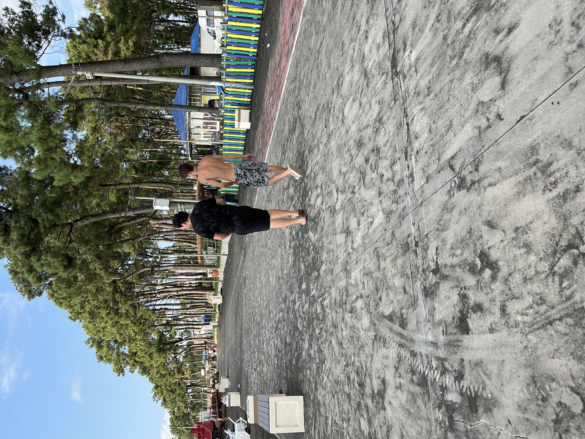Two young men walk away from the camera on a sandy promenade in Ureki, Georgia. The man on the left wears a black t-shirt and black shorts. The man on the right wears a black and white patterned swim trunks. They are walking along a paved path lined with trees on the left and a colorful fence on the right. The promenade appears to be at a beach resort, with lounge chairs and some buildings visible in the background. The sky is clear and blue.