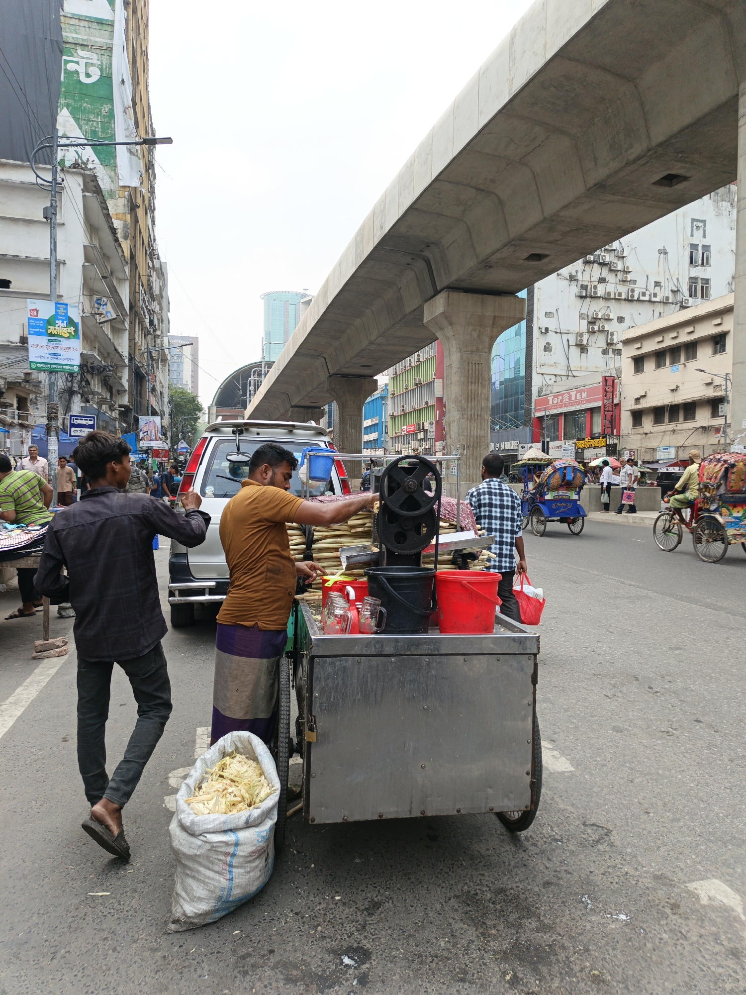 The image captures a street scene in Dhaka, Bangladesh, with a focus on a sugarcane juice vendor operating from a metal cart on a busy street. The vendor, wearing a brown shirt and a lungi, is actively preparing the juice. A young man in a dark shirt stands to the left, potentially a customer. A large bag filled with sugarcane remnants sits next to the cart.
Behind the vendor, a white SUV is parked, seemingly obstructing traffic. The street is filled with other vehicles, including cycle rickshaws, and pedestrians. An elevated concrete structure, likely part of the Dhaka metro rail system, dominates the skyline.
Buildings line both sides of the street, displaying a mix of architectural styles. The street is cluttered with various signs and banners, typical of a bustling urban environment. The weather appears overcast, contributing to the relatively subdued lighting of the scene.