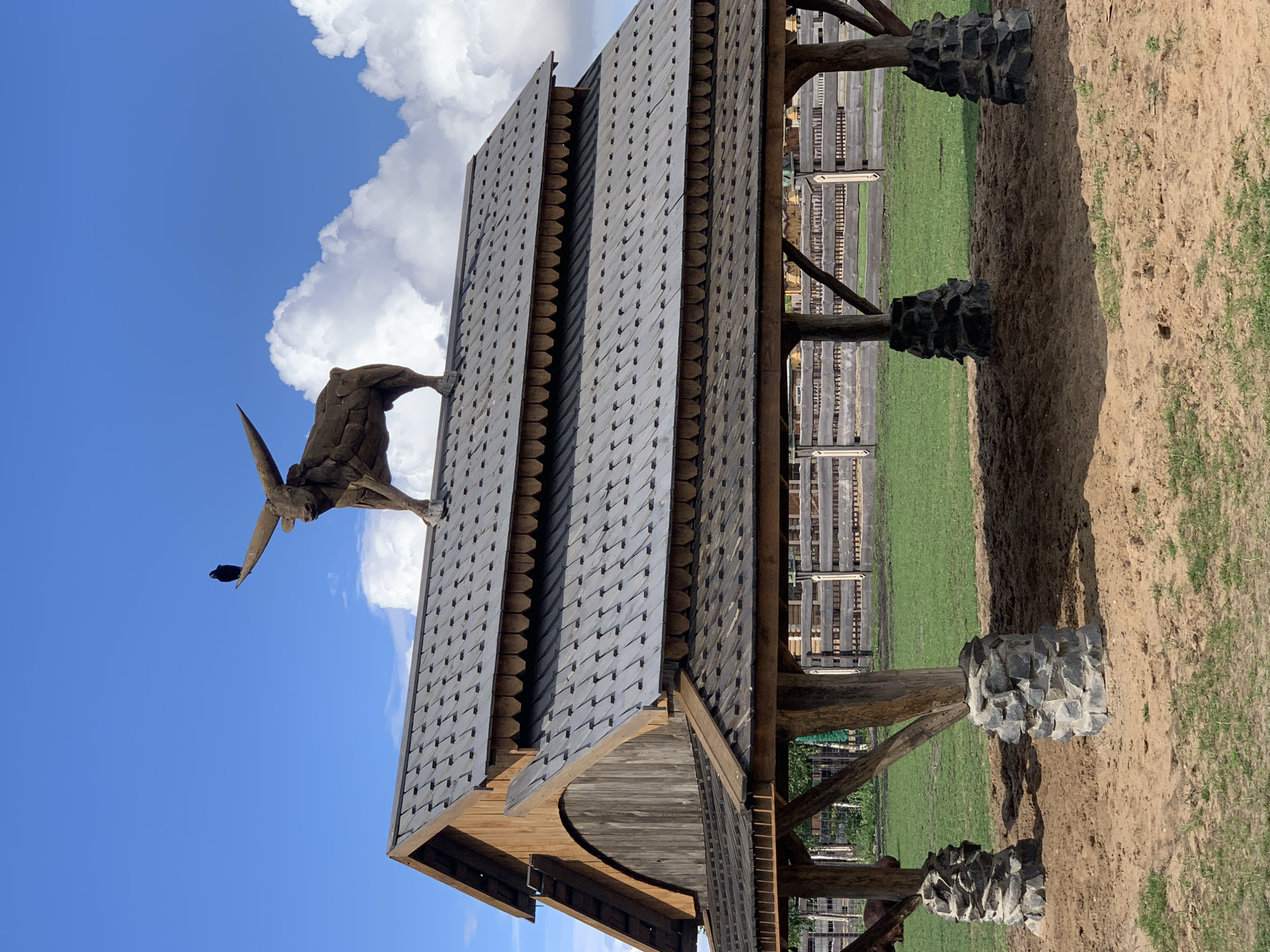 The image shows a wooden pavilion in a rural outdoor setting. The pavilion has a tiered, shingled roof and is supported by rough-hewn wooden posts resting on stone bases. Atop the highest point of the roof stands a large, sculpted bull figure, seemingly made of metal or wood, with prominent horns. Perched on one of the bull's horns is a small black bird, possibly a crow or raven. The ground around the pavilion is sandy with patches of dry grass. In the background, a wooden fence encloses a green grassy area, suggesting a farm or park environment. The sky above is a vibrant blue with scattered white cumulus clouds. The location is identified as Malakhovetsky Selsoviet, Belarus. There are no people visible in the image.