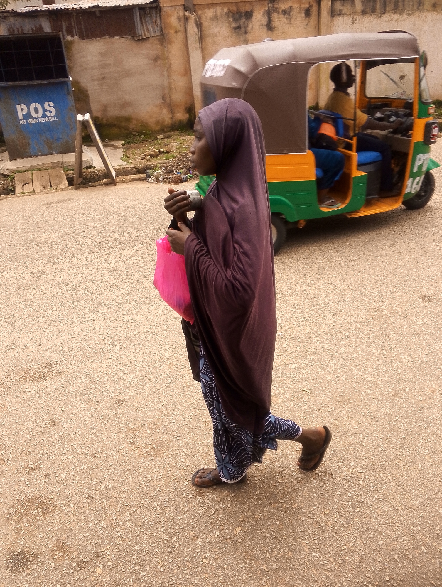 A young woman wearing a purple hijab and a patterned blue and white skirt walks across a paved road in front of a brightly colored auto-rickshaw. She is carrying a pink plastic bag in her right hand. The auto-rickshaw, which has a green, yellow, and brown canopy, is occupied by a person driving. In the background, there is a building with a corrugated metal roof and a blue dumpster with the text "POS PAY YOUR NEPA BILL" visible. The environment appears to be an urban street.