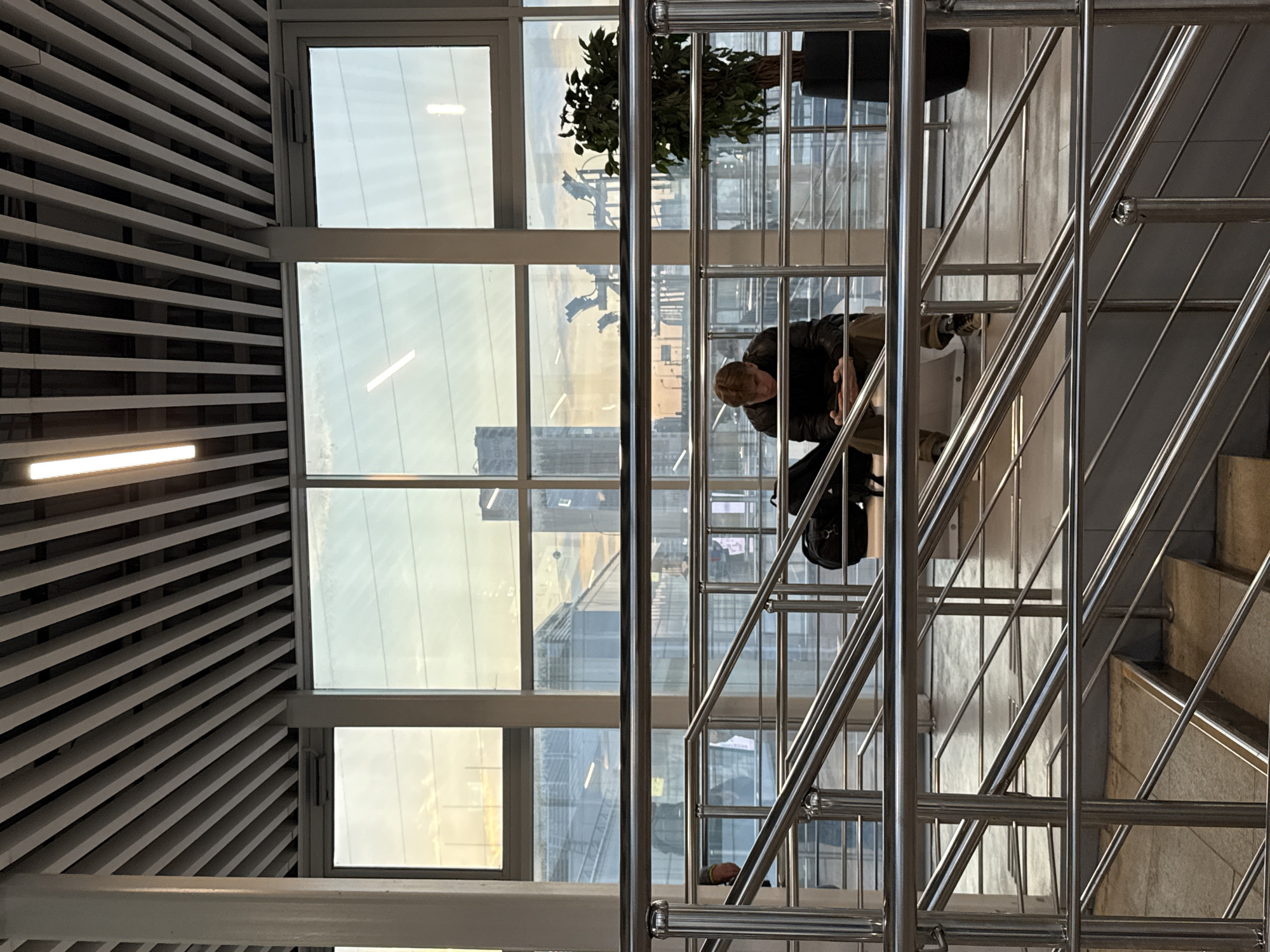 Here is a detailed description of the image:
**Setting:**
The image appears to be taken indoors, possibly in a modern office building or public space. A large window is visible in the background, offering a view of a city. There are horizontal slat blinds on the upper left of the image and a metal staircase with railings is prominent in the foreground.
**Objects:**
- **Window:** A large window with a view of a city, possibly Moscow, as indicated in the prompt. There are distinct office buildings in the background.
- **Metal Staircase:** A set of stairs with a metallic frame and railings.
- **Plant:** A potted plant is visible near the upper part of the frame by the window.
- **Backpack:** A backpack is hanging from the handrail of the staircase
**Subjects and People:**
- **Individual on Stairs:** A person is on the staircase, appearing to be going down. The individual is wearing a dark jacket and lighter-colored trousers.
**Environment:**
- **Lighting:** The lighting seems to be a mix of natural light from the window and artificial indoor lighting.
- **Architecture:** Modern architectural elements are visible, including the large window and the metallic staircase.
**Actions:**
- An individual is going down the staircase.