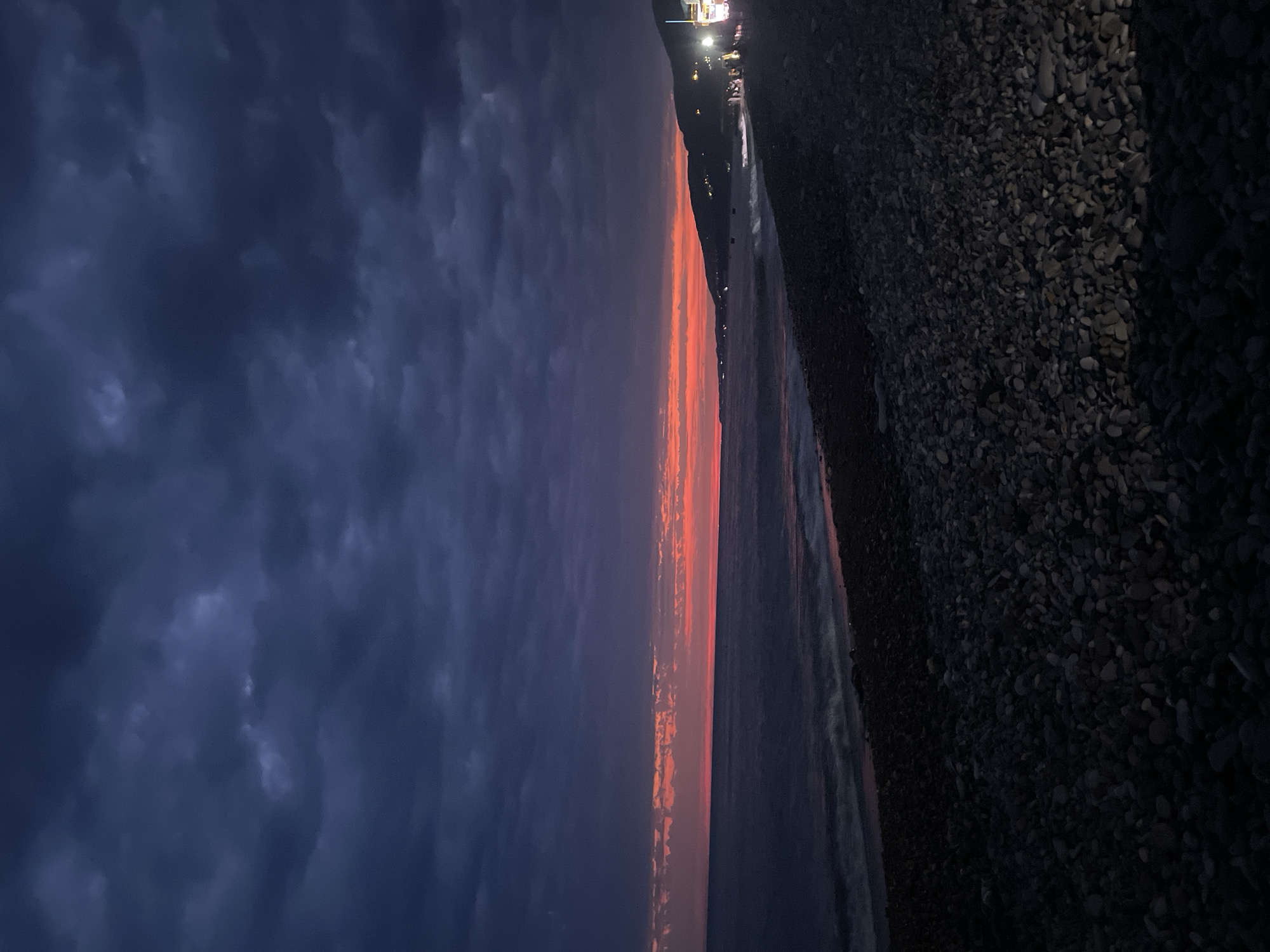 An eye-level, full shot captures a tranquil seascape at dusk. The sky, a canvas of deep indigo, is streaked with dramatic, dark clouds that transition to a vibrant band of orange and red near the horizon. Below, the sea mirrors the moody sky, with gentle waves lapping at a dark, pebbled beach. The shoreline curves into the distance, where a hilly landscape is dotted with lights, suggesting a settlement. A few figures are visible on the beach, gathered near illuminated structures. The overall impression is one of serene natural beauty and subtle human presence against a dramatic twilight backdrop.