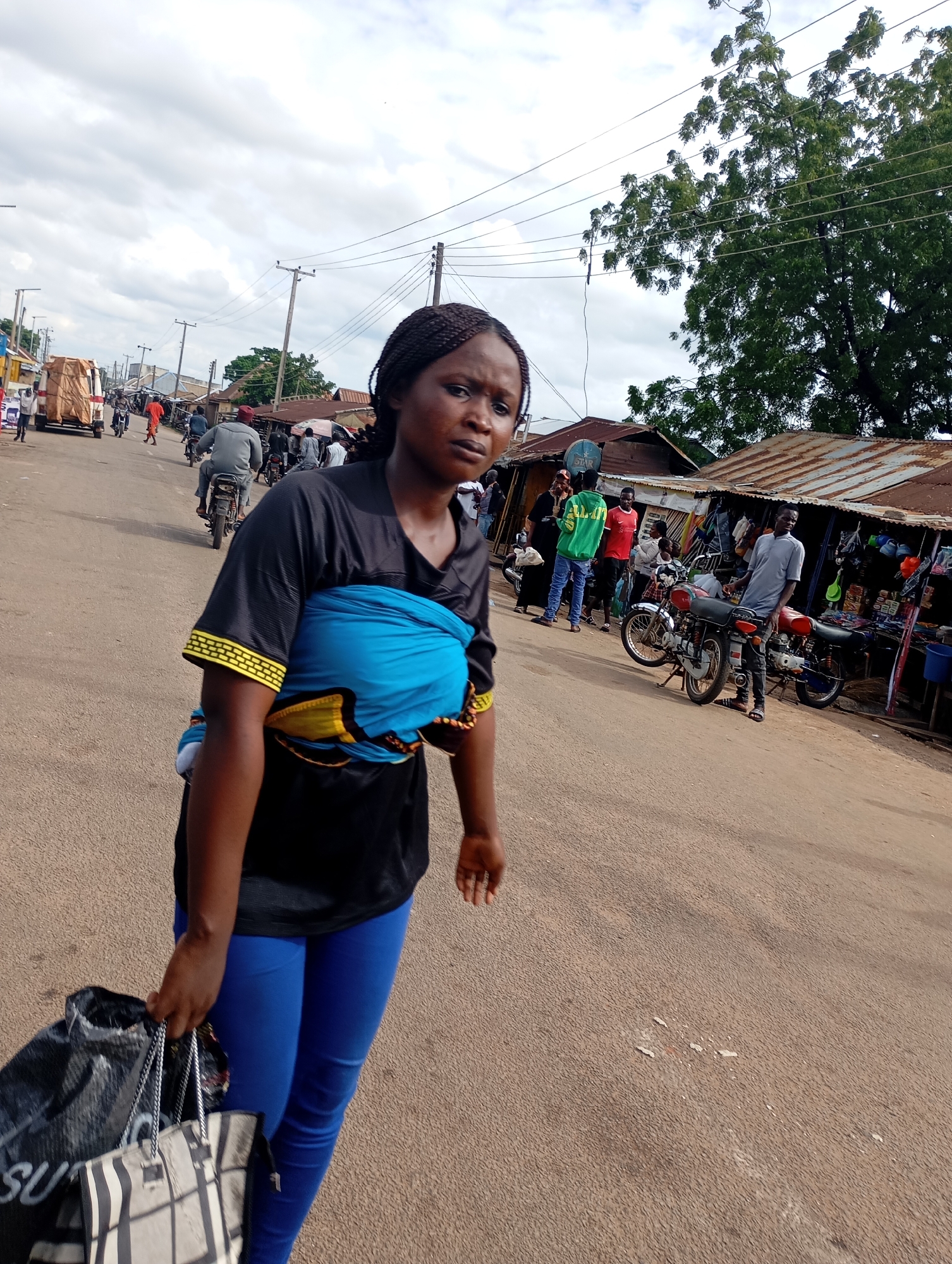 In the city of Shendam, Nigeria, a Black woman with braided hair walks towards the viewer on a paved road lined with market stalls and trees. She wears a black t-shirt with yellow trim on the sleeves and bright blue leggings. A vibrant blue cloth is wrapped around her chest, holding an unseen infant. She carries two bags in her left hand, one black and the other a patterned tote. In the background, several people are visible, some on motorcycles and others walking near the stalls, which display various goods under corrugated metal roofs. Power lines crisscross the sky, and the day appears overcast.