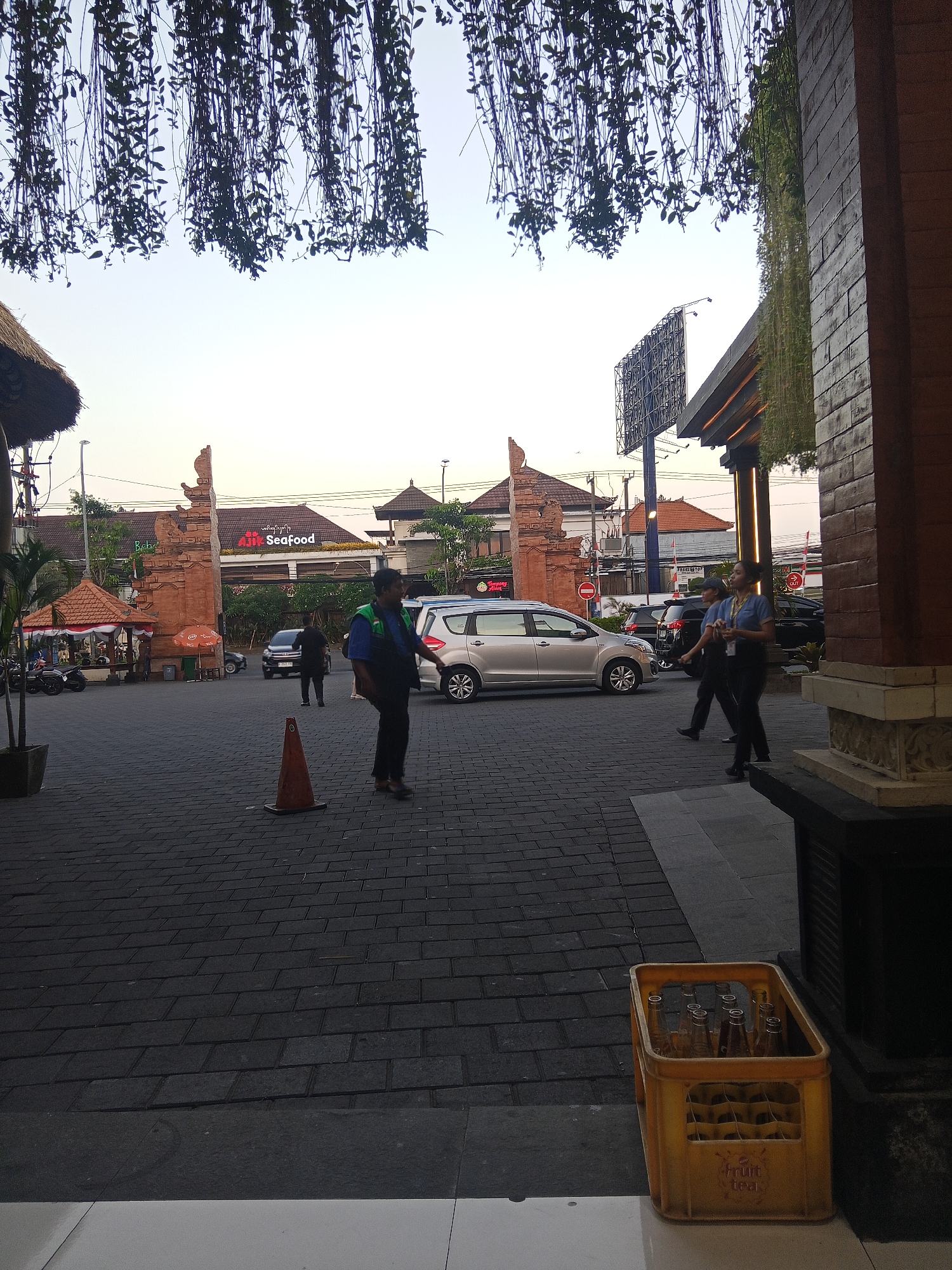 In Kuta, Indonesia, this outdoor scene captures a paved area with people and vehicles. Dominating the background is a red structure with arched openings and traditional Balinese architectural elements, likely an entrance to a complex or building. A sign above reads "Air Seafood," indicating a restaurant. Several vehicles, including a silver minivan and two black SUVs, are parked or moving in the background. In the foreground, a man in a blue vest and dark pants stands in the center of the paved area. To the right, a yellow plastic crate filled with empty glass bottles sits near a brick wall. The sky is clear and appears to be either early morning or late evening. Lush green vines hang from above, creating a canopy over the scene.