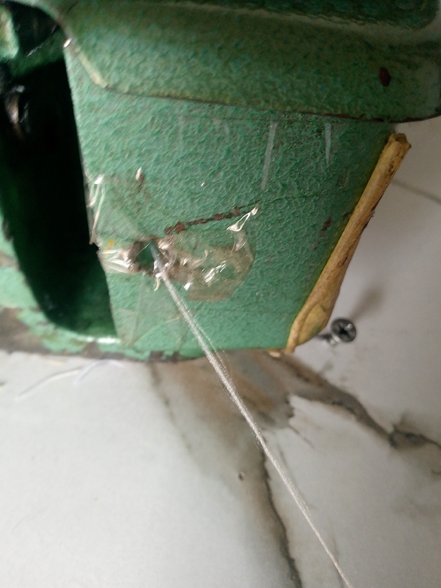 The image shows a close-up view of a green metal object, possibly part of a machine. A thin white thread is running through a small hole in the object, secured with clear tape. A piece of dried straw or wood is leaning against the object. In the background, a screw is visible in a marbled surface. The scene appears to be indoors, with the lighting suggesting it is daytime. The image does not provide any specific information or visual cues to indicate its location within Jauro Yinu, Nigeria.