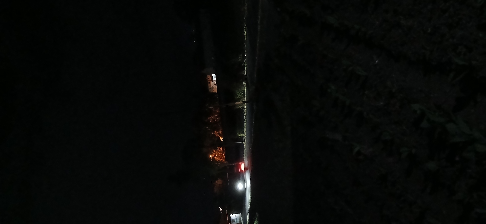 The image depicts a dimly lit outdoor scene at night, likely in a rural or suburban area of Sekarputih, Indonesia, characterized by minimal light sources and the presence of vegetation. In the middle ground, a vehicle, possibly a car or van, is visible with its headlights illuminated, casting a bright glow on the road ahead. The taillights of the vehicle are also visible, indicating its movement.
To the left of the vehicle, a building with some illuminated windows is partially visible. In the background, across the road, there are more buildings and trees, with some faint orange light emanating from behind the trees, possibly from streetlights or another building. The foreground and much of the midground are dark and largely obscured by shadows, with patches of vegetation visible. The overall impression is of a quiet night scene on a road with some surrounding structures and natural elements.