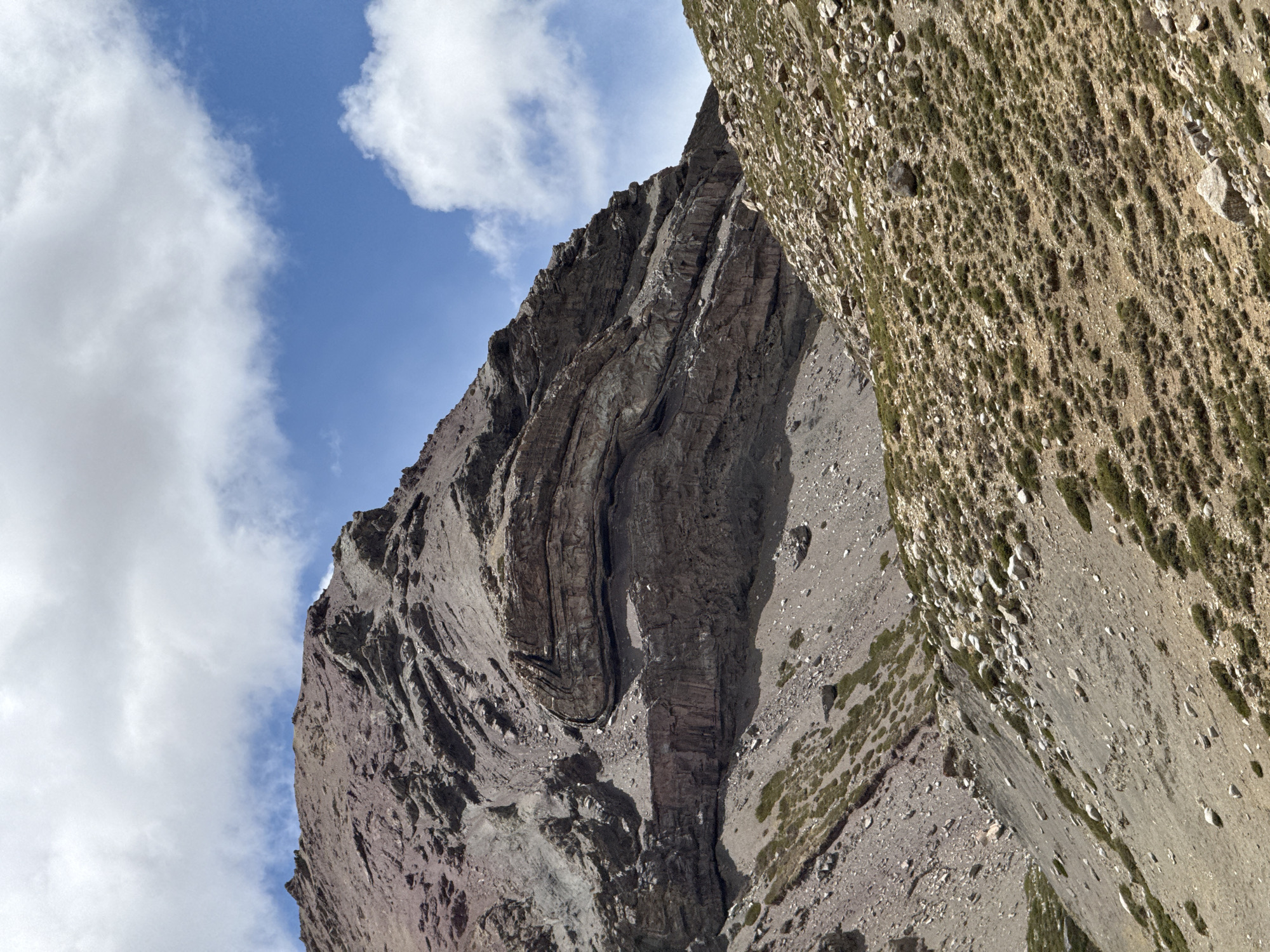 The image, oriented vertically, depicts a rugged mountain landscape under a partly cloudy blue sky. A massive, dark-colored rock face dominates the left and central parts of the frame, showcasing prominent geological folds where distinct layers of rock are bent and curved. These intricate patterns of folded strata suggest significant geological activity. Sunlight illuminates sections of this cliff, creating contrasting shadows that accentuate its textured surface and deep crevices.
To the right of the central rock formation, a steep slope rises, covered with a mix of scree, loose rocks, and sparse green vegetation. This terrain appears harsh and arid. The sky occupies the upper portion of the image, featuring a vibrant blue adorned with several fluffy white cumulus clouds.
The scene conveys a sense of wild, untouched nature and geological grandeur, captured during what appears to be late morning or early afternoon given the bright lighting and shadows. No people, man-made structures, or visible text are present. The provided location context for this mountain scene is Parga town, China.