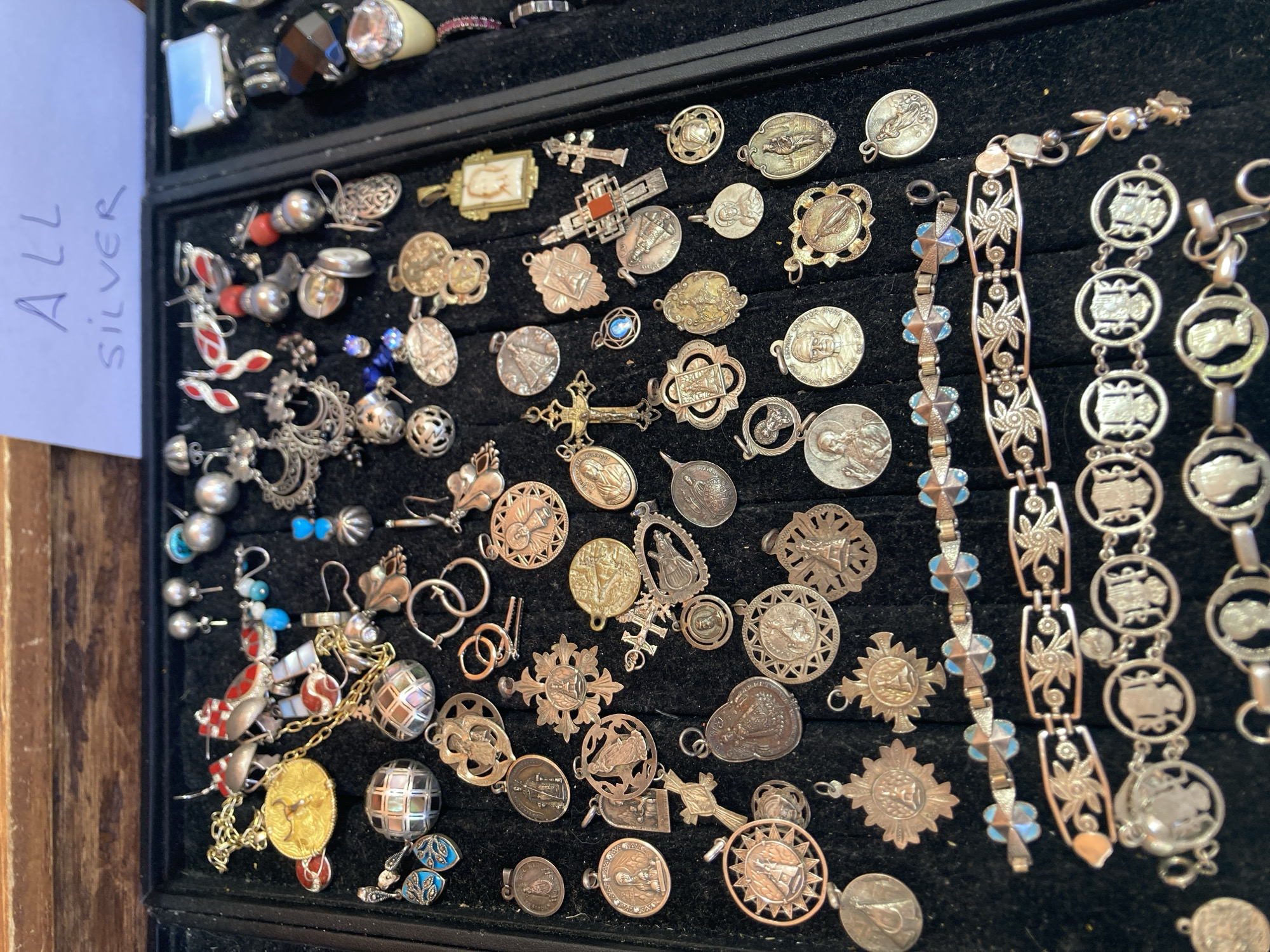 The image shows a close-up, overhead view of a display of assorted jewelry, primarily consisting of pendants and bracelets. The jewelry is laid out on a dark, felt-like surface, possibly within a display case. Various pendants are visible, including many circular or oval religious medallions featuring figures like the Virgin Mary and saints, as well as crosses. There are also several bracelets, some with intricate floral or decorative links, and others featuring repeating portrait motifs. A note handwritten on a piece of paper in the upper right corner reads "ALL SILVER." The background suggests a rustic setting, possibly a market stall or antique shop, with a wooden surface visible at the top of the frame. No people are visible in the image. The context of Barcelona, Spain is not deducible from the visual information provided in the image itself.