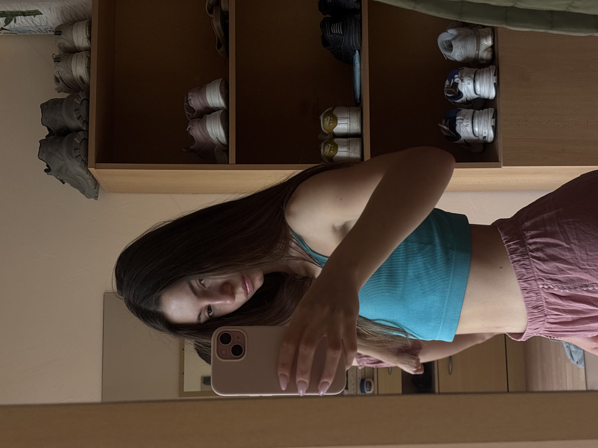 A young woman with long, dark brown hair is taking a mirror selfie in a dimly lit room. She is wearing a turquoise ribbed crop top and pink linen shorts. Her left hand is holding a pink smartphone, and her right arm is bent at the elbow. In the background, there is a tall wooden shelf unit filled with various pairs of sneakers. To the left of the woman is a large mirror reflecting the room. Part of a chest of drawers with two handles is visible on the left side of the frame. The scene appears to be within a residential setting. There is no visible indication of the city of Varna, Bulgaria.
