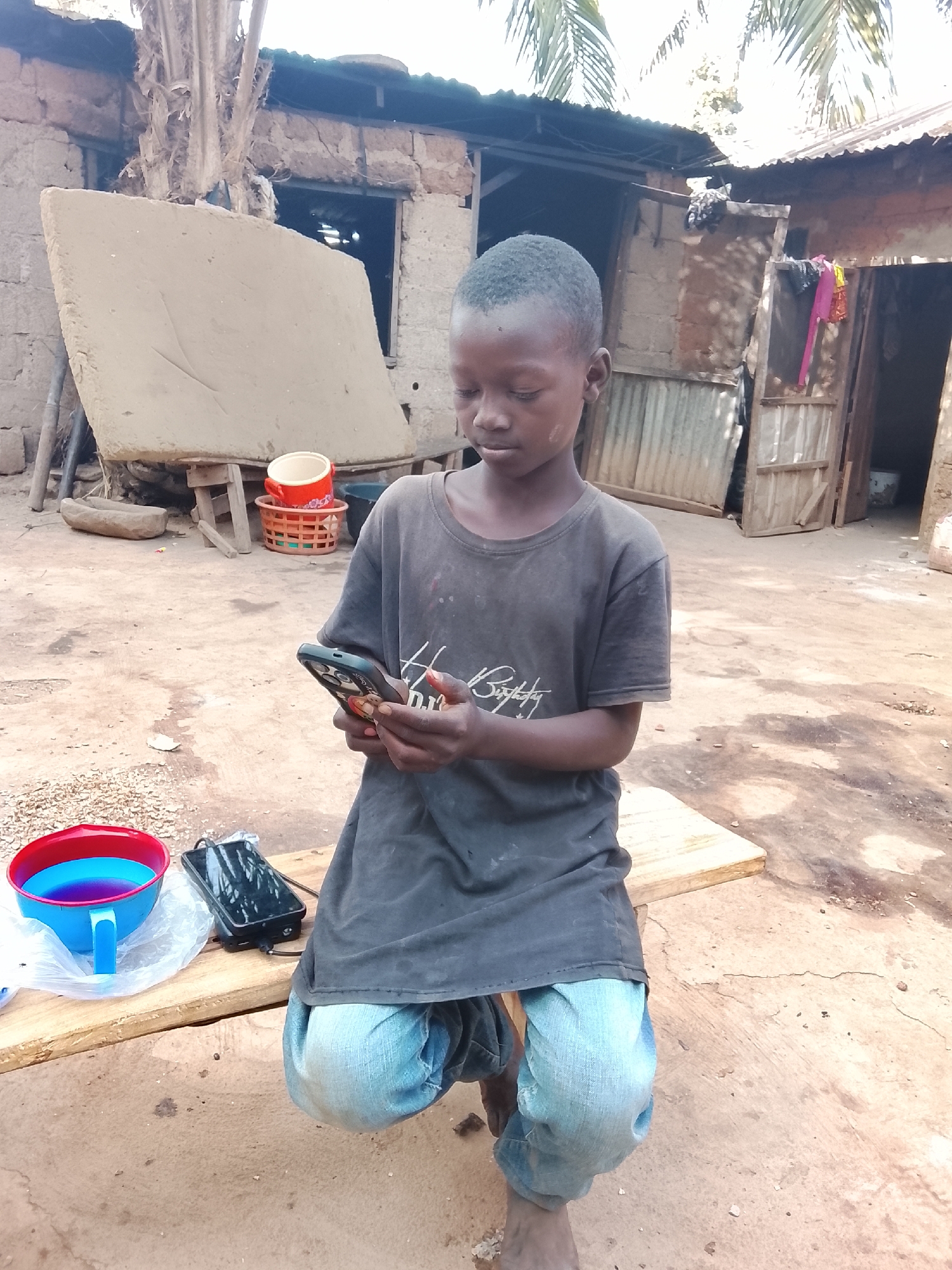 A young boy sits on a wooden plank outdoors, holding a smartphone in his hands, his gaze focused on the screen. He is dressed in a dark T-shirt and light-blue jeans. Next to him on the plank sits a plastic bowl with blue liquid and a smartphone connected to a power bank. The setting appears to be a courtyard or backyard with earthen ground, surrounded by buildings made of mud and corrugated iron. There are signs of poverty. The location is in the city of Unknown, Unknown.