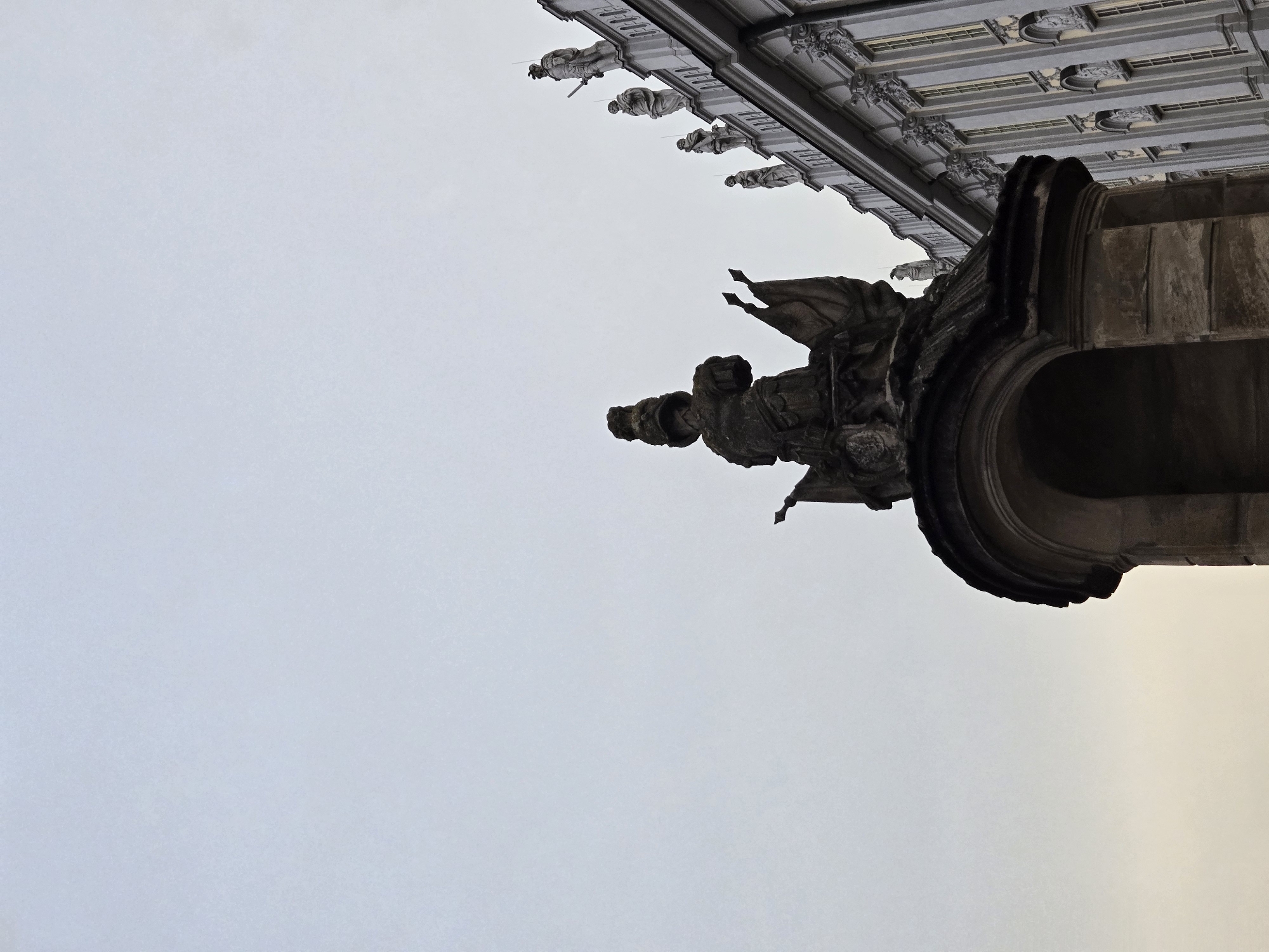 This image is an upward-angle shot of the exterior of a building with architectural details, specifically gargoyles and ornate carvings. In the foreground, a large, dark, stone gargoyle is prominently featured. It appears weathered and menacing, with sharp angles and a gaping mouth. Behind and above it, a series of smaller, lighter-colored figures, also possibly gargoyles or statues, line the facade of the building. These figures are more detailed, with what appear to be human-like forms dressed in robes, some holding objects that could be swords or staffs. The building itself is grand, with a patterned ceiling or frieze visible above the figures. The sky is overcast and pale, suggesting it could be daytime with cloudy weather. The overall impression is one of ancient architecture, possibly a cathedral or a historic public building. The orientation of the image is unconventional, with the building appearing to hang from the top of the frame. No text is visible in the image. The location context is Ansbach, Germany, which aligns with the Gothic or Neo-Gothic architectural style suggested by the gargoyles and ornate stonework.