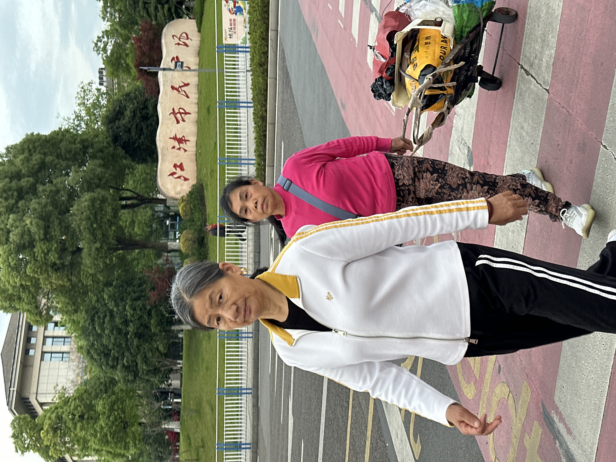 This image captures an outdoor scene in Jiangjin, China, featuring two women in what appears to be a pedestrian area or sidewalk. The day is overcast, suggesting daytime with diffused lighting.
In the foreground, an older woman with gray hair wears a white zip-up jacket with yellow trim and black athletic pants with white stripes. She has a neutral expression and looks directly at the camera. A small, indistinct logo, possibly "Hjp," is visible on her jacket.
Behind her, another woman, dressed in a bright pink long-sleeved top and dark patterned trousers, is engaged in pulling a two-wheeled utility cart or trolley. The cart is heavily loaded with various bags and bundled items, potentially recyclables. One yellow bag in the cart displays partial text, including "OUR AN" and some Asian characters. This woman also looks towards the camera with a neutral demeanor.
To the left of the women, a prominent stone sign is visible. It bears large red Chinese characters which read "江津市民场" (Jiāngjīn Shìmínchǎng), translating to "Jiangjin Citizen's Square" or "Jiangjin People's Square," clearly indicating the location.
The ground features distinct red and white painted stripes, suggesting a pedestrian crossing or a designated pathway. In the background, there are lush green trees, a grassy area, a white fence, and parts of modern buildings under an overcast sky. The scene depicts a common urban or park setting with everyday activities.