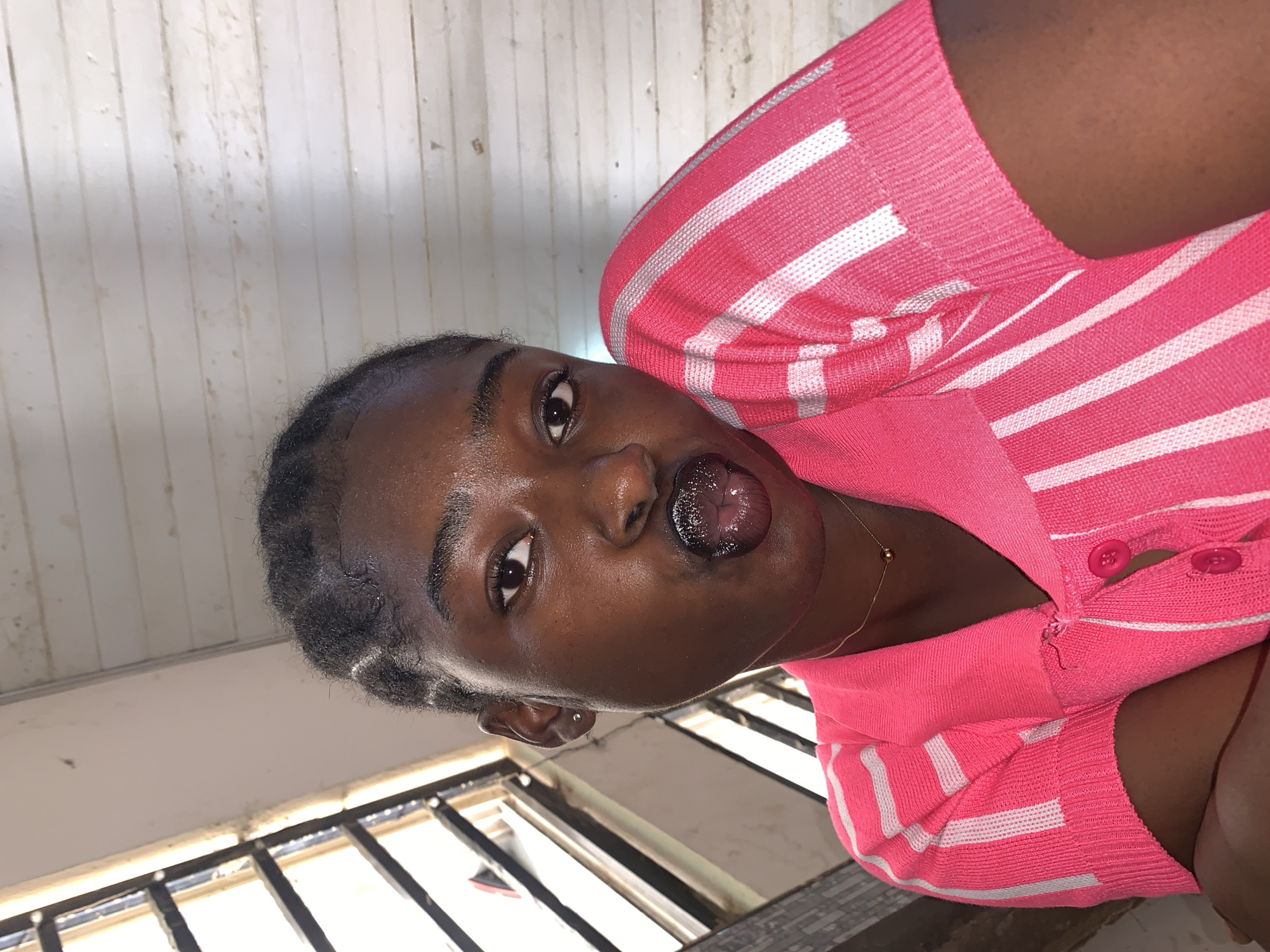 A dark-skinned woman is photographed from a low angle, resulting in a distorted perspective of the surrounding environment. She is wearing a pink, striped sweater. Her mouth is open, her tongue protruding slightly. A gold necklace is visible around her neck. The background includes a window with bars and a white wall made of what appears to be wooden planks. The location is Kabong, Nigeria.