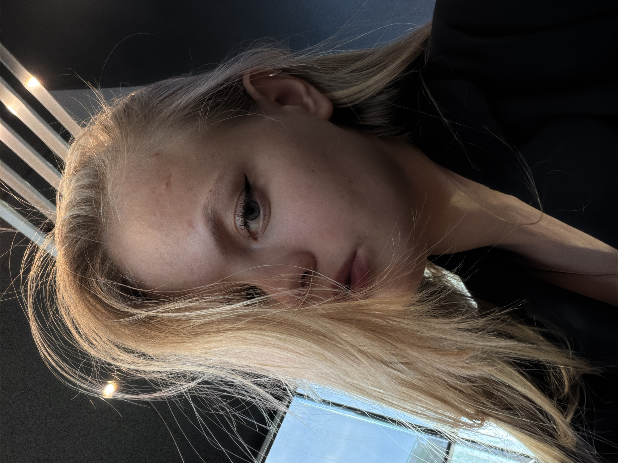 A close-up portrait of a young woman with blonde hair. Her head is tilted, partially obscuring her face with her hair. She has light skin, light eyes, and minimal makeup. She is wearing a black top. The background is blurred, showing a window with white slats and dark surroundings, potentially an interior setting. The image appears to have been taken indoors under artificial lighting. No location information is provided or discernible from the image.