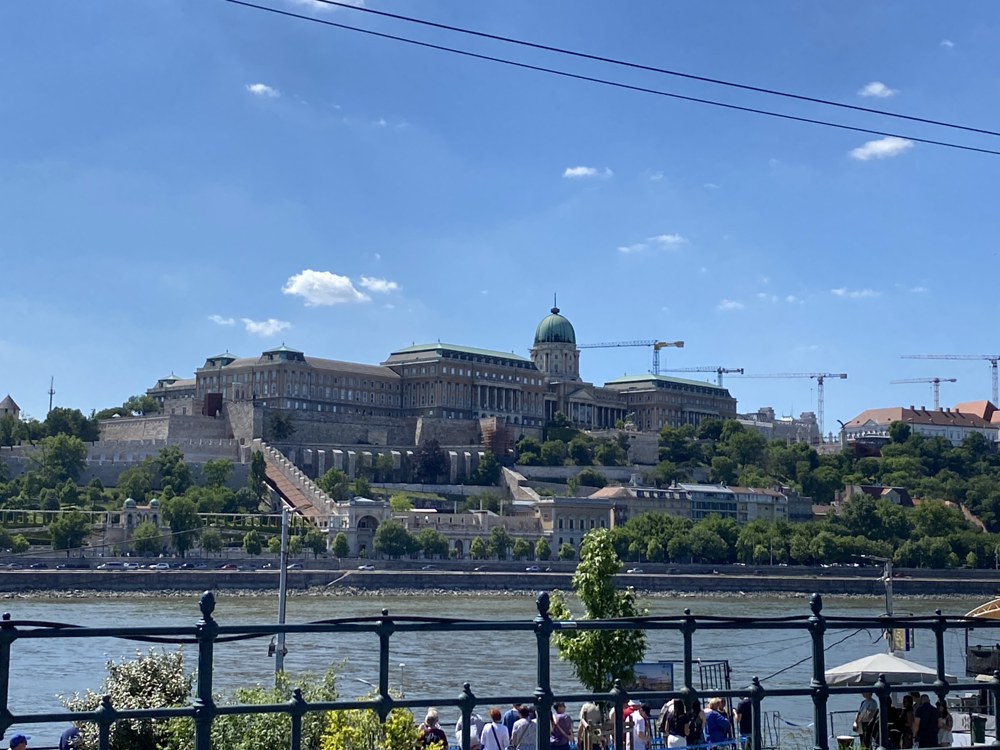 The image captures a daytime view of Buda Castle in Budapest, Hungary, seen from across the Danube River. The castle complex sits atop Castle Hill, with its Baroque architecture dominating the landscape. Several construction cranes are visible behind the castle, indicating ongoing work in the area. The Danube flows in the foreground, with a dark metal fence and shrubbery lining the riverbank. A small group of people stands behind the fence. The sky is blue with scattered clouds, and two overhead wires stretch across the top of the frame. The scene suggests a tourist location with renovation work underway at the historic castle.