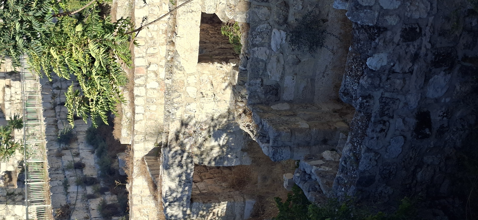 The image depicts an archaeological site in Jerusalem, Israel, likely part of the ancient city walls or a related structure, characterized by weathered stone construction and overgrown vegetation. In the foreground, irregular stone blocks form partial walls and debris, suggesting a state of ruin or excavation. Two arched openings, possibly doorways or passageways, are visible in a more intact section of stone walling further back, with dry grass and shadows within them. Above these openings, terraces are built from similar stone blocks, with sparse, dry vegetation growing in the crevices. A metal railing with green vertical bars runs along the top of the uppermost stone wall, indicating a preserved section or a protective barrier. A branch of a leafy green tree extends into the upper right portion of the frame, its shadow cast upon the stone structures. The overall scene is one of historical excavation and natural overgrowth in an urban archaeological setting.