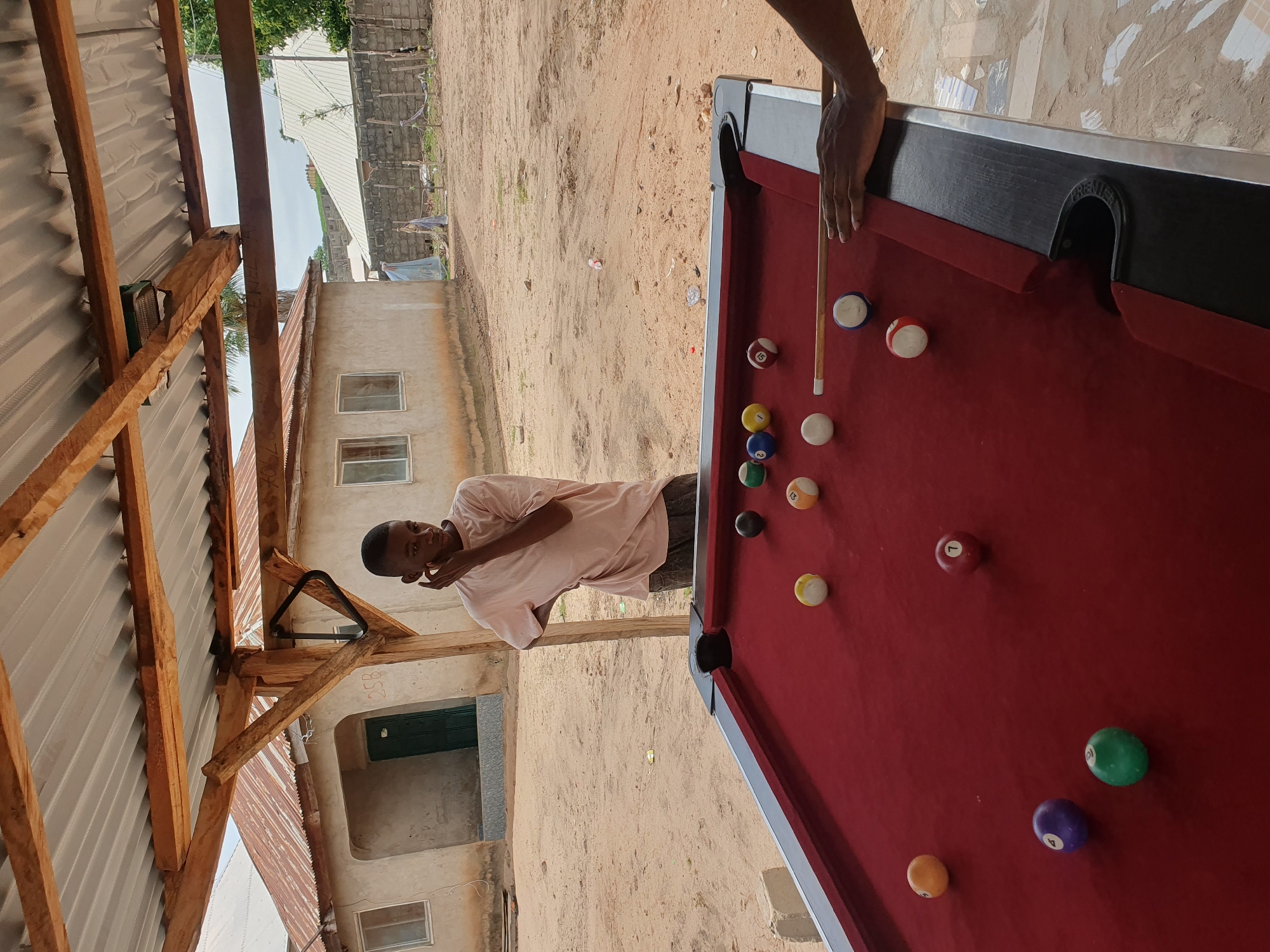 A young Black male, wearing a light pink t-shirt and dark pants, stands under a rustic wooden shelter with a corrugated metal roof, leaning his left arm on a wooden beam and touching his chin with his right hand. He is looking down at a red billiard table. On the table, there are several billiard balls of various colors and numbers, including a white cue ball. A person's hand is visible on the right side of the frame, positioned as if about to strike the cue ball with a cue stick. The ground around the table is sandy. In the background, there is a light-colored, single-story building with a rusty corrugated metal roof and windows, and further in the distance, other buildings and a brick wall are visible. The scene appears to be in an outdoor, informal setting, potentially in Lokoja, Nigeria, given the architectural style and environment.