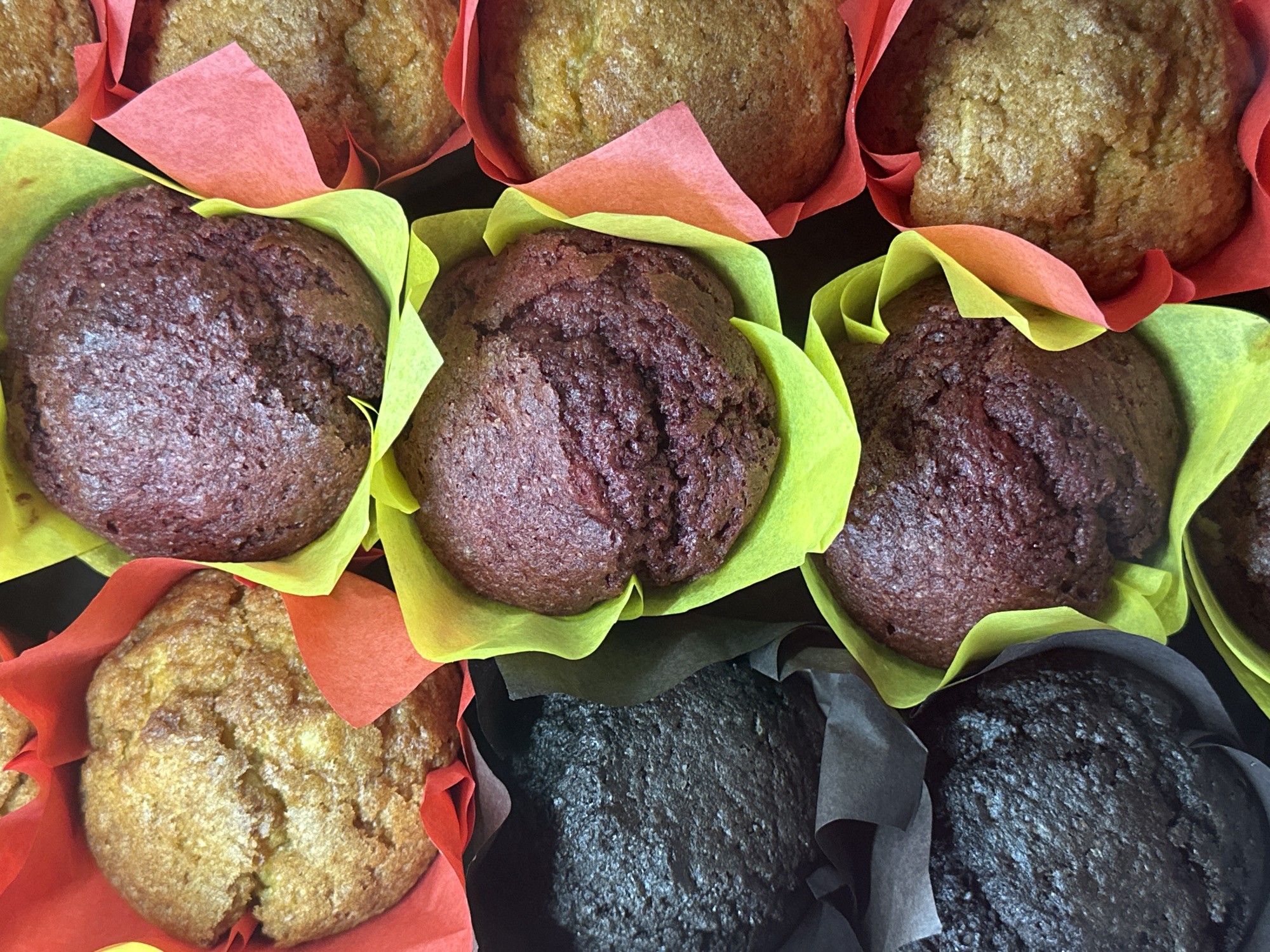 The image provides a close-up, top-down view of multiple muffins arranged closely together. There are several distinct types of muffins, easily identified by their color and the various paper liners they are presented in.
In the upper section, golden-brown muffins, likely a plain, vanilla, or fruit-based variety, are visible, mostly enclosed in vibrant red and orange paper liners. Below these, a row of darker, reddish-brown muffins, possibly red velvet or berry-flavored, are prominently displayed in bright yellow and lime green liners. Towards the bottom of the frame, very dark brown or black muffins, suggesting a chocolate or double chocolate variety, are nestled in dark grey or black paper liners.
The muffins feature a textured, slightly uneven baked top surface. Their arrangement suggests they are part of a display, such as in a bakery, cafe, or at a catered event. No people or specific activities are visible. The lighting is bright and even, highlighting the colors and textures of the baked goods. There are no discernible location cues, time of day, or visible text within the image.