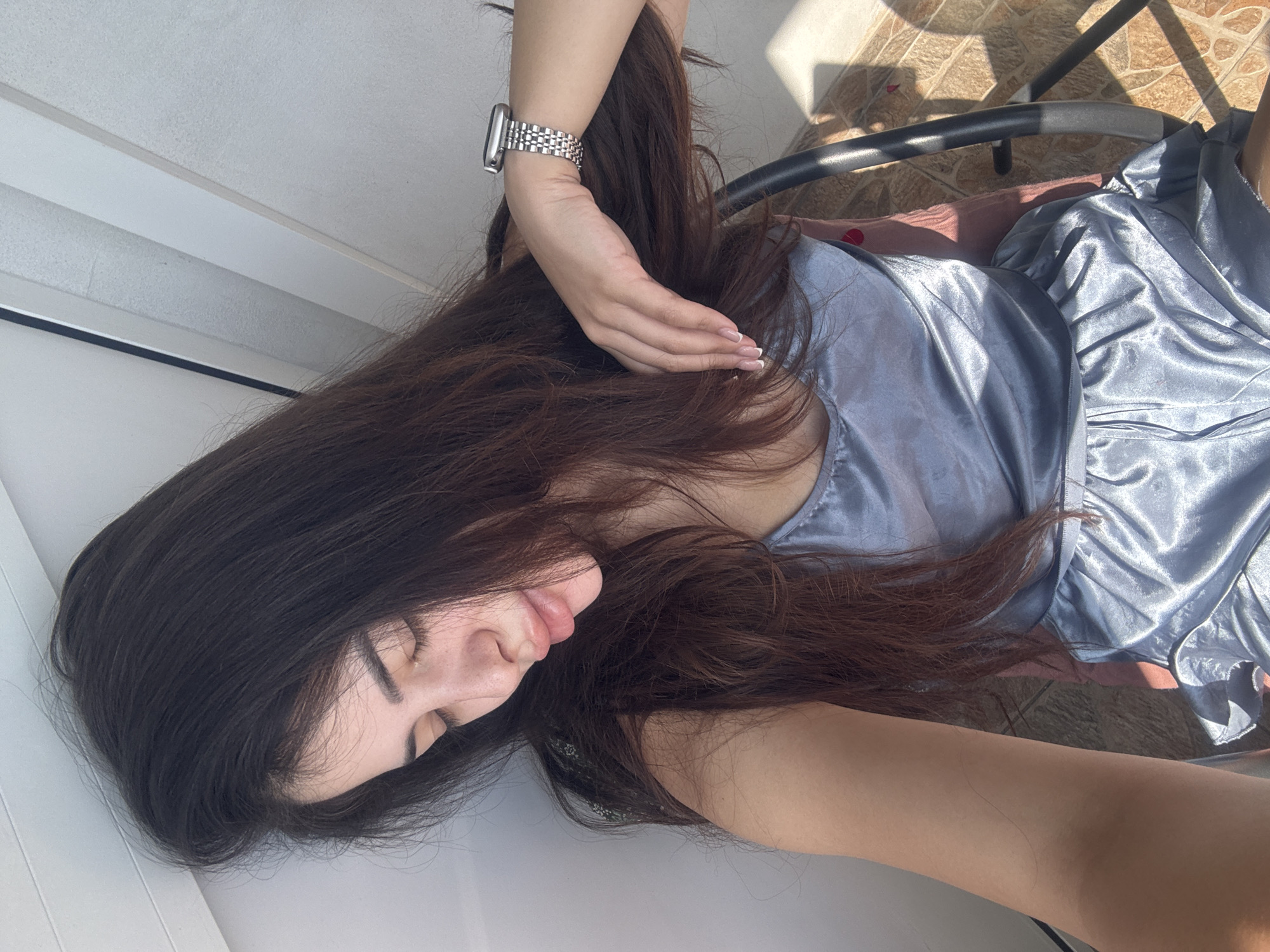 This media file, captured in Plovdiv, Bulgaria, features a young woman in a relaxed and peaceful state. She has long, dark brown hair that cascades around her as she lies down or leans back, with her eyes gently closed and a serene expression. She is dressed in a light blue or silvery-grey satiny garment, which catches the sunlight and creates a lustrous texture.
On her left wrist, she wears a silver-colored watch with a metallic link band. Her left hand, adorned with neatly manicured nails featuring a French tip design, is subtly placed within her hair.
The scene is brightly illuminated by natural light, indicated by the strong, distinct shadows cast across the environment. The background suggests an outdoor or semi-outdoor setting, possibly a balcony or patio, featuring a tiled floor with a warm, earthy tone and a geometric pattern. A black-framed chair with a visible pink cushion is also present. A plain white surface, likely a wall or part of a ceiling, forms the backdrop in the upper left. The intense sunlight points to either morning or late afternoon. No text is visible in the image.