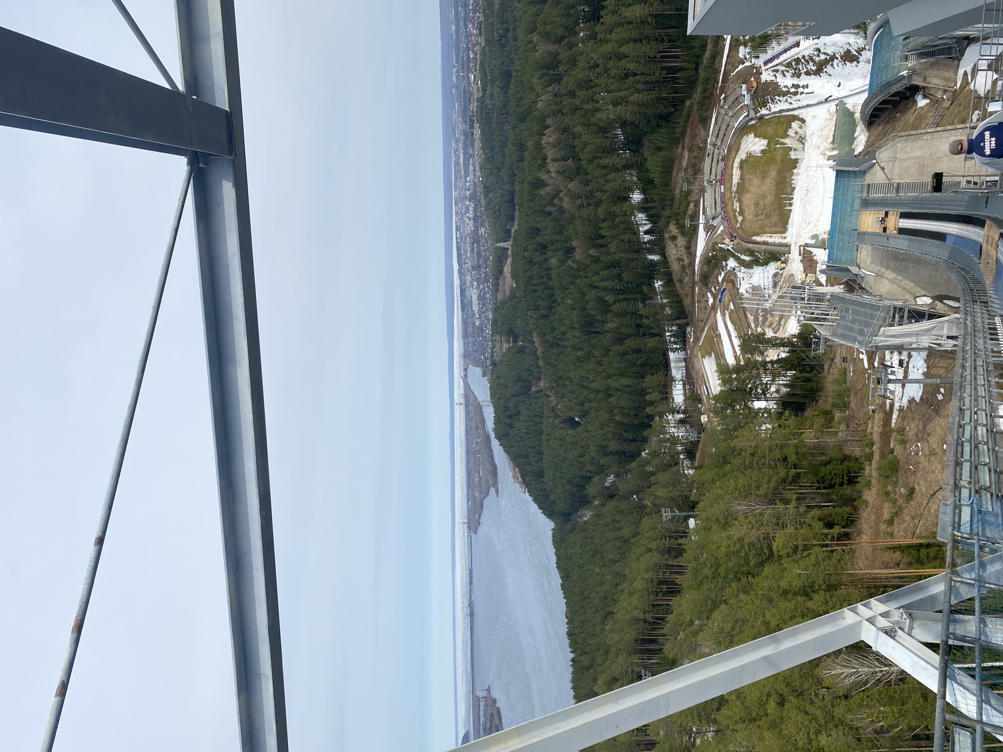 View of the Lysva River from the ski jumping complex in Прикамский, Russia. There are several pine trees on the river's edge. The ski jumping complex has a green field at the end of the ski jump surrounded by spectator stands. The metal structures from the complex's tower are in the foreground.