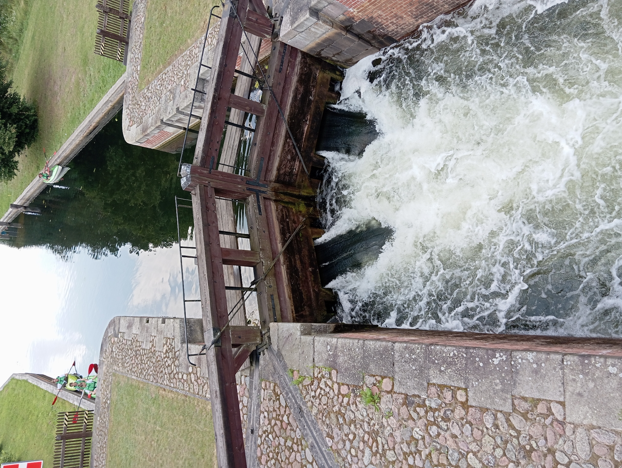In Mikaszówka, Poland, a canal lock is in operation, with water cascading forcefully from two sluices into a lower section of the waterway. The lock gate is made of weathered brown wood, with metal reinforcements and cables visible. The surrounding banks are lined with stone and cobblestones, and a grassy area with a wooden fence is present to the left. Two people in kayaks, wearing life vests and holding paddles, are positioned in the upper section of the canal, to the left of the lock mechanism. One kayak is directly behind the other, and the individuals appear to be waiting. The water is a deep green, reflecting the sky and surrounding trees.