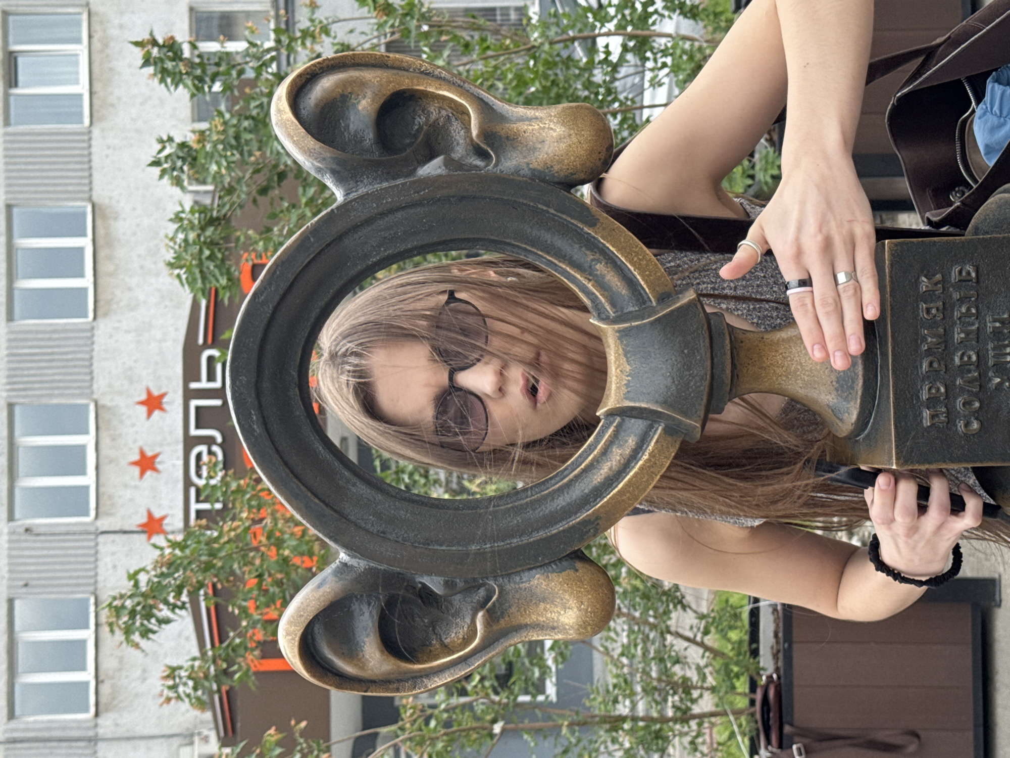 A young woman with long brown hair and sunglasses stands behind a bronze sculpture of a large ear. The sculpture is part of a public art installation, with the base inscribed in Russian. The woman is looking forward and touching the sculpture with her hands. The background shows a building with windows and a sign that reads "ОТЕЛЬ" (HOTEL), indicating the location is near a hotel. Green trees and shrubs are visible in the foreground and background. The overall scene is outdoors, likely in a city environment in Perm, Russia.