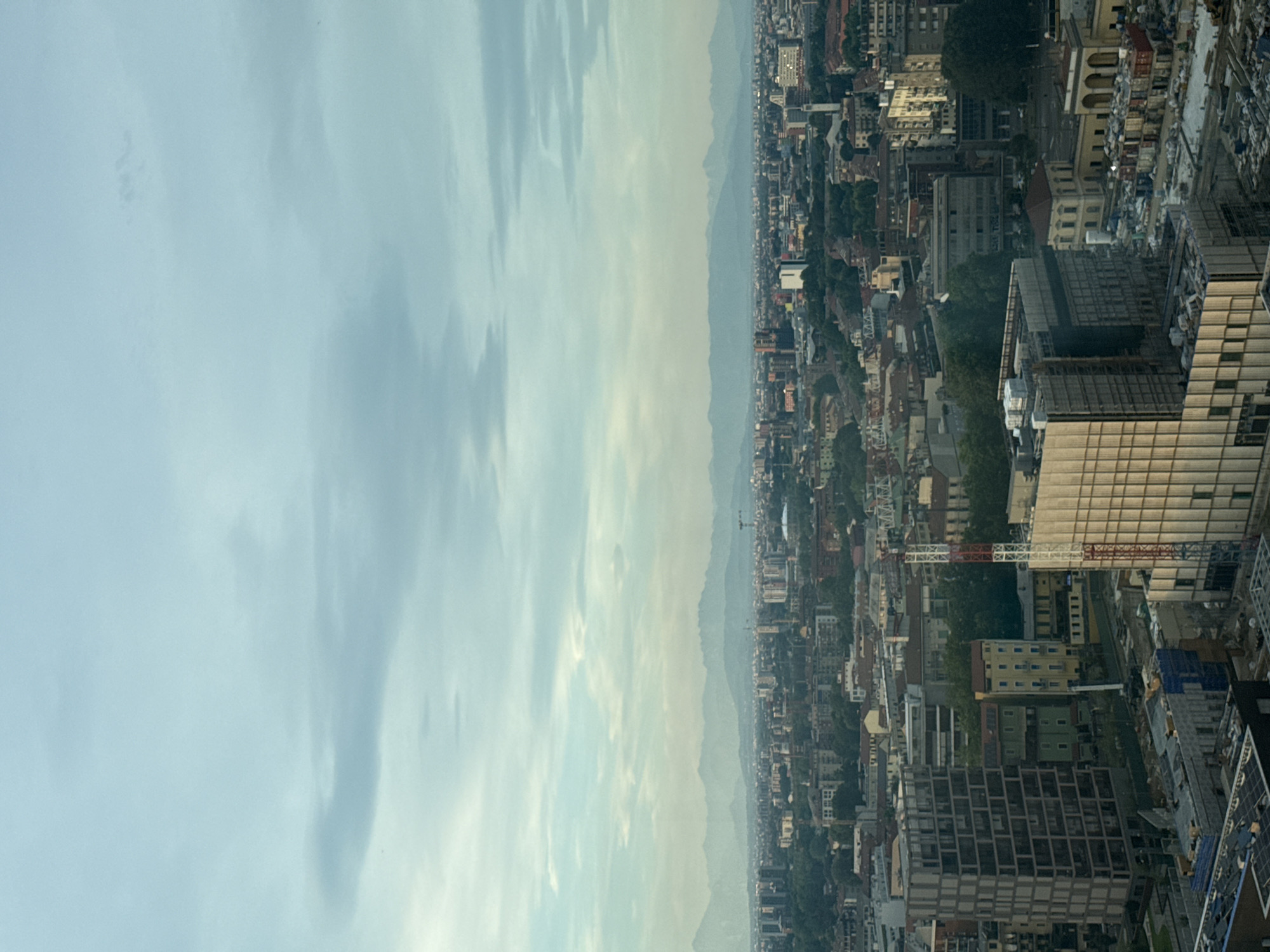 This is an aerial, high-angle, vertical shot of the city of Milan, Italy. The sky is overcast with soft, muted light, casting a gentle haze over the urban landscape. In the foreground, a large, modern building with a grid-like facade is visible. Adjacent to it, another contemporary structure with a ribbed, metallic exterior is under construction, with scaffolding and materials present on its lower levels. Further into the mid-ground, a dense cityscape unfolds with a mix of residential and commercial buildings, many featuring reddish-brown tiled roofs. Trees are interspersed throughout the urban fabric, adding touches of green. A tall, red and white construction crane stands prominently near the center. In the distance, a range of hazy mountains forms the horizon. The overall scene depicts a sprawling European city with a blend of old and new architecture under a diffused sky.