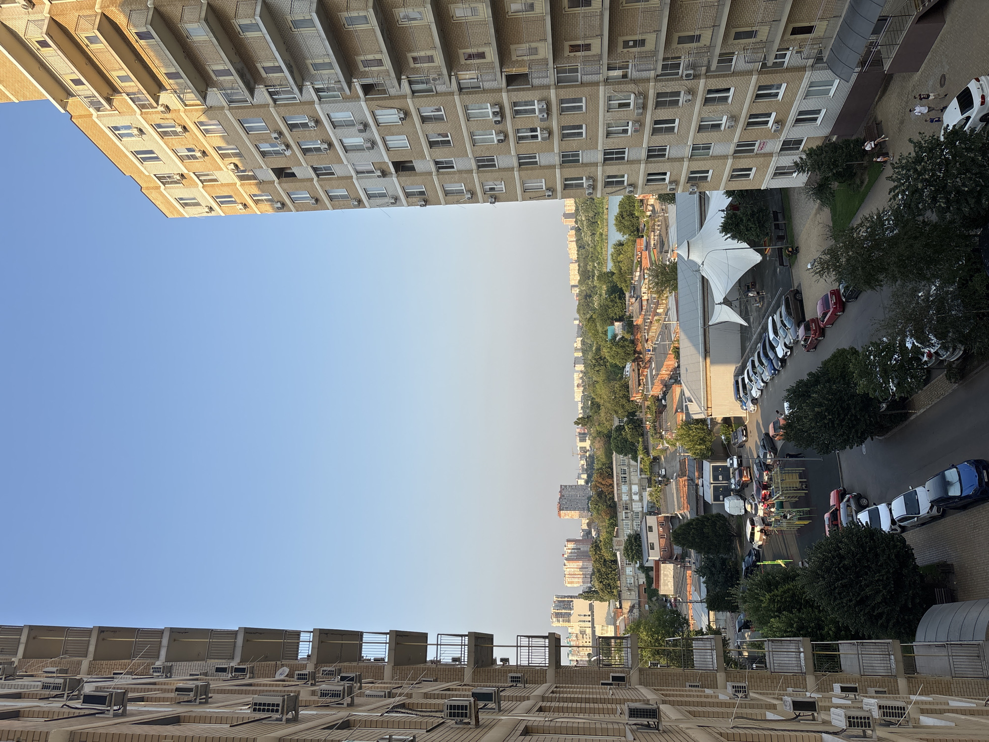 The image captures a view from a high-rise apartment building in Krasnodar, Russia, looking out over a residential area and a distant body of water. Two multi-story apartment buildings flank the foreground, with numerous air conditioning units visible on their brick facades. Below, a street lined with parked cars winds through a neighborhood featuring smaller buildings, garages, a playground, and trees. In the distance, a hazy skyline of more apartment buildings and a large body of water can be seen under a clear blue sky. A white, tent-like structure is also visible in the middle ground. Several individuals are walking and sitting on benches in the lower right portion of the image.