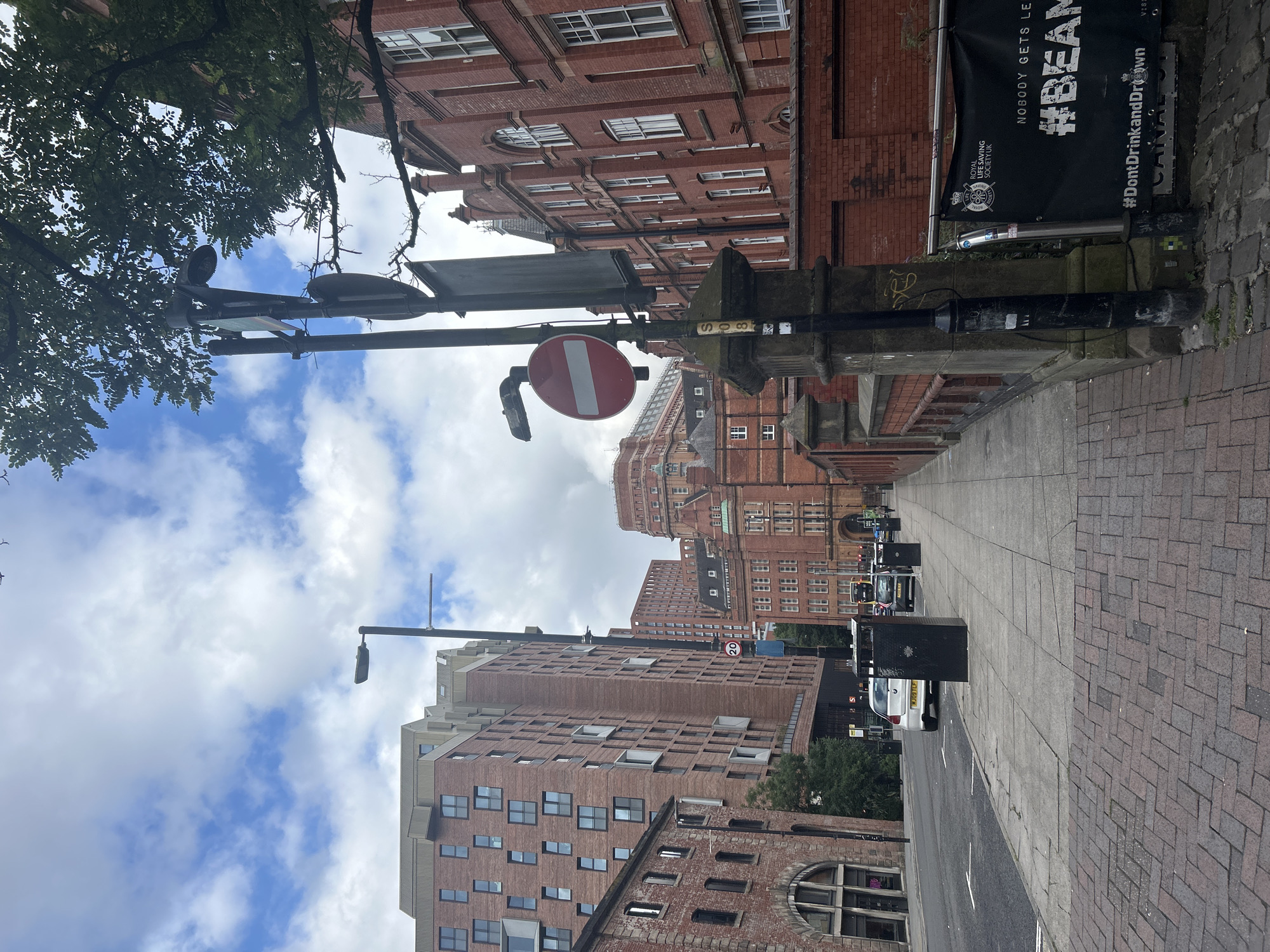 A medium shot captures a street scene in Manchester, United Kingdom, with a focus on a sidewalk and a row of brick buildings. The foreground is dominated by a red and gray patterned brick sidewalk. To the left of the sidewalk is a road with a white car and two black trash bins. The sidewalk is lined with a brick wall and a series of lampposts.
Behind the wall, on the right, a large red brick building with arched windows spans across the frame. Further back, a modern, multi-story red brick building with numerous windows stands prominently. The sky above is partly cloudy with scattered white clouds against a blue backdrop.
In the mid-ground, several street signs are visible, including a red circular sign with a white horizontal bar, indicating "no entry" or "do not enter." A tall black lamppost with multiple fixtures and a banner that reads "ROYAL LIFE SAVING SOCIETY UK NOBODY GETS LEFT #BEAM #DontDrinkandDrown" is also present. The street appears to be in an urban setting, likely a commercial or residential area.