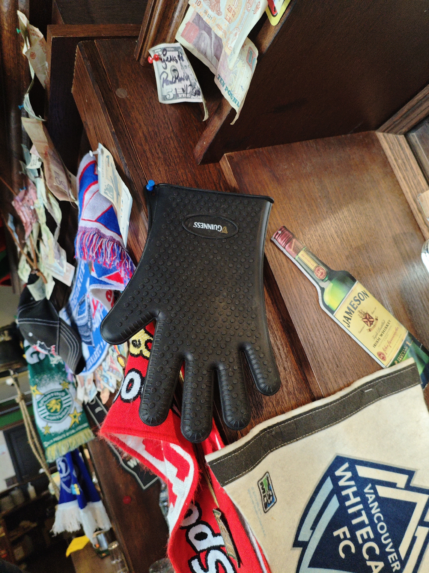 The image displays a close-up view of a collection of items attached to a wooden structure, possibly a wall or shelf in a bar or pub. The items include several banknotes, some of which are pinned with thumbtacks. Also visible are multiple scarves, a black oven mitt with the Guinness logo, and a cutout image of a Jameson Irish Whiskey bottle. At the bottom right, a cream-colored banner with the text "VANCOUVER FC" is partially visible. The lighting suggests an indoor setting. There is no visible indication of the city of Kyiv, Ukraine, or any specific location.
