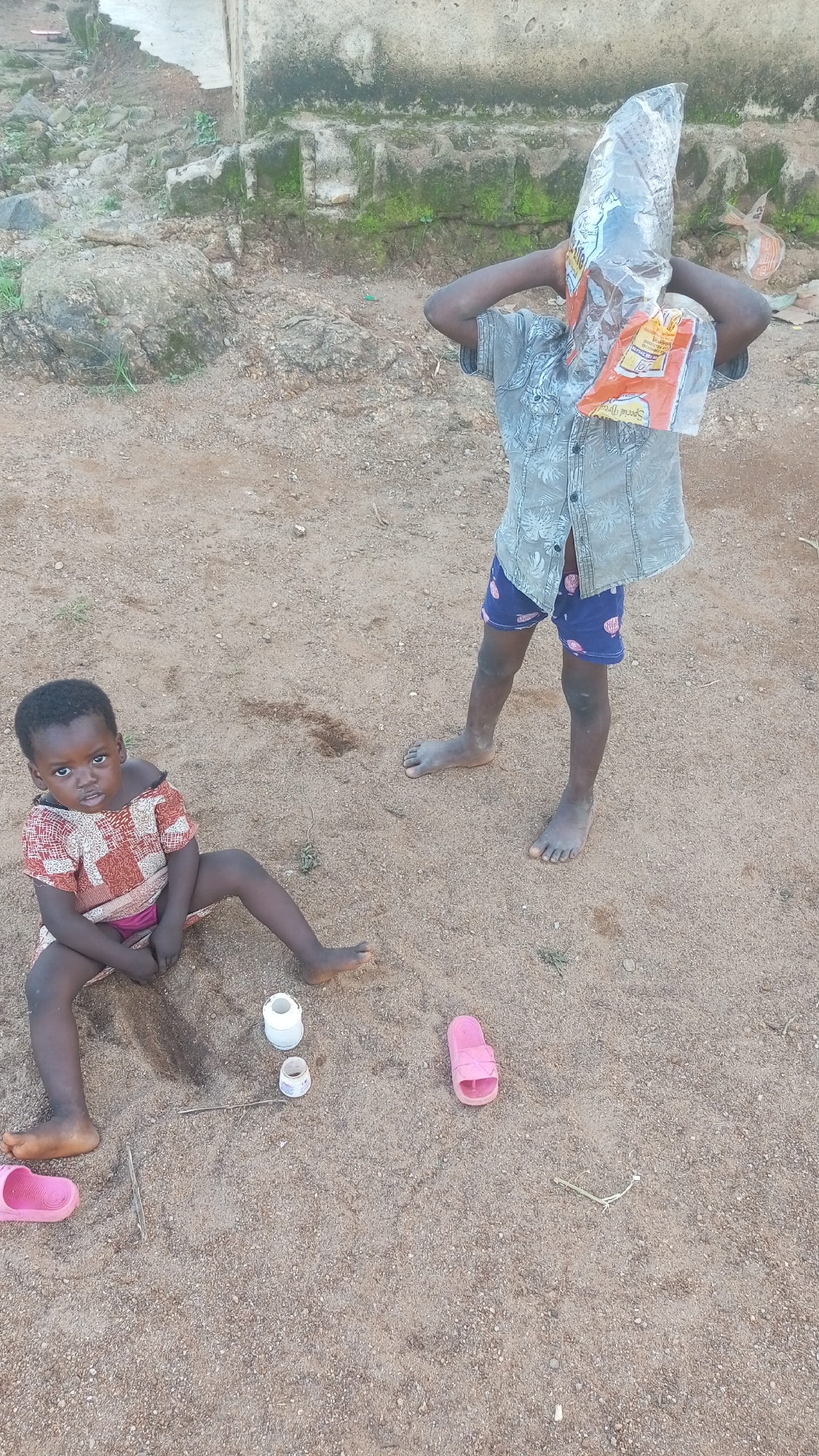 The image depicts two young children in an outdoor, sandy setting, likely in Kagarko South, Nigeria. In the foreground, a young girl with dark skin sits on the sand, facing the camera with a neutral expression. She is wearing a patterned, off-the-shoulder dress and a pink garment underneath. To her left, a pink sandal lies on the ground. In the mid-ground, a second child, possibly a boy, stands with their back to the camera, holding a clear plastic bag over their head, obscuring their face. This child is wearing a patterned short-sleeved shirt and blue shorts with a pattern. A pink sandal is also visible near this child. Scattered on the sand between the children are two small white containers and a thin stick. The background shows a rough, uneven surface with rocks and some vegetation, leading to a weathered, light-colored structure. The lighting suggests it is daytime.