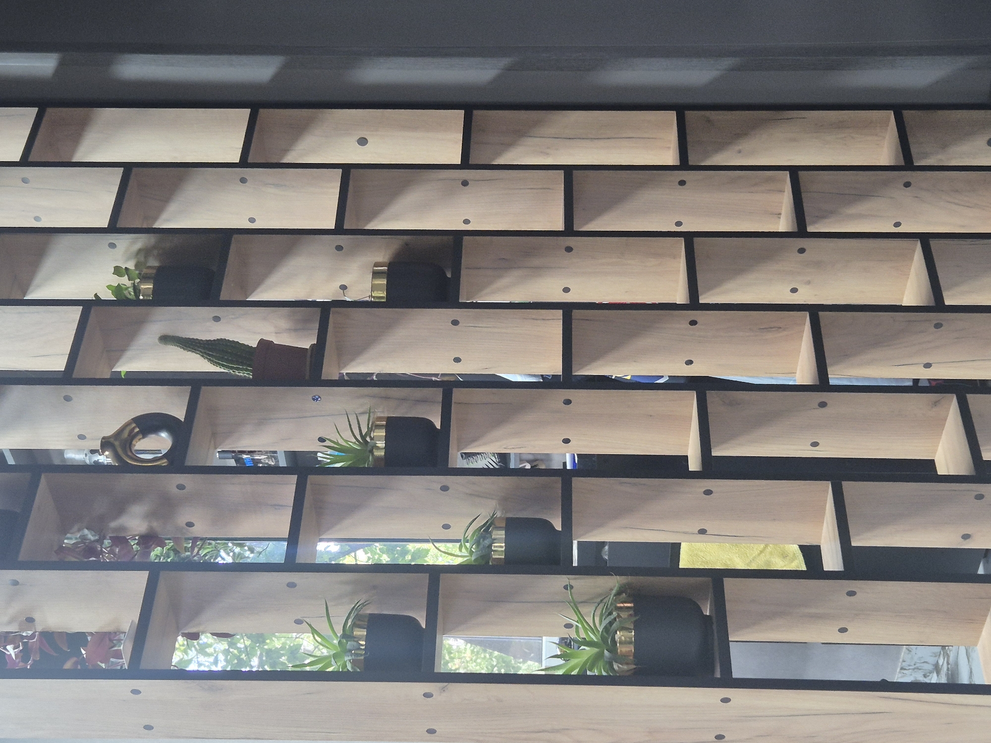 The image displays a modern, open-shelf wooden room divider with black metal framing. The divider is segmented into numerous cubbies of varying heights and widths, some of which are empty, while others contain decorative items. These items include potted plants, a decorative vase with a gold and dark brown finish, and potentially other home decor items that are partially visible or out of focus. The divider is placed in an indoor setting, with natural light filtering through from behind some of the cubbies, suggesting it is positioned near a window or an opening. The wall on the right side of the image is painted a dark gray color. There are no people visible in the image. The scene is static and appears to be a still life composition within an interior space. The location in the city of Kokshetau, Kazakhstan is not discernable from the visual information provided in the image.