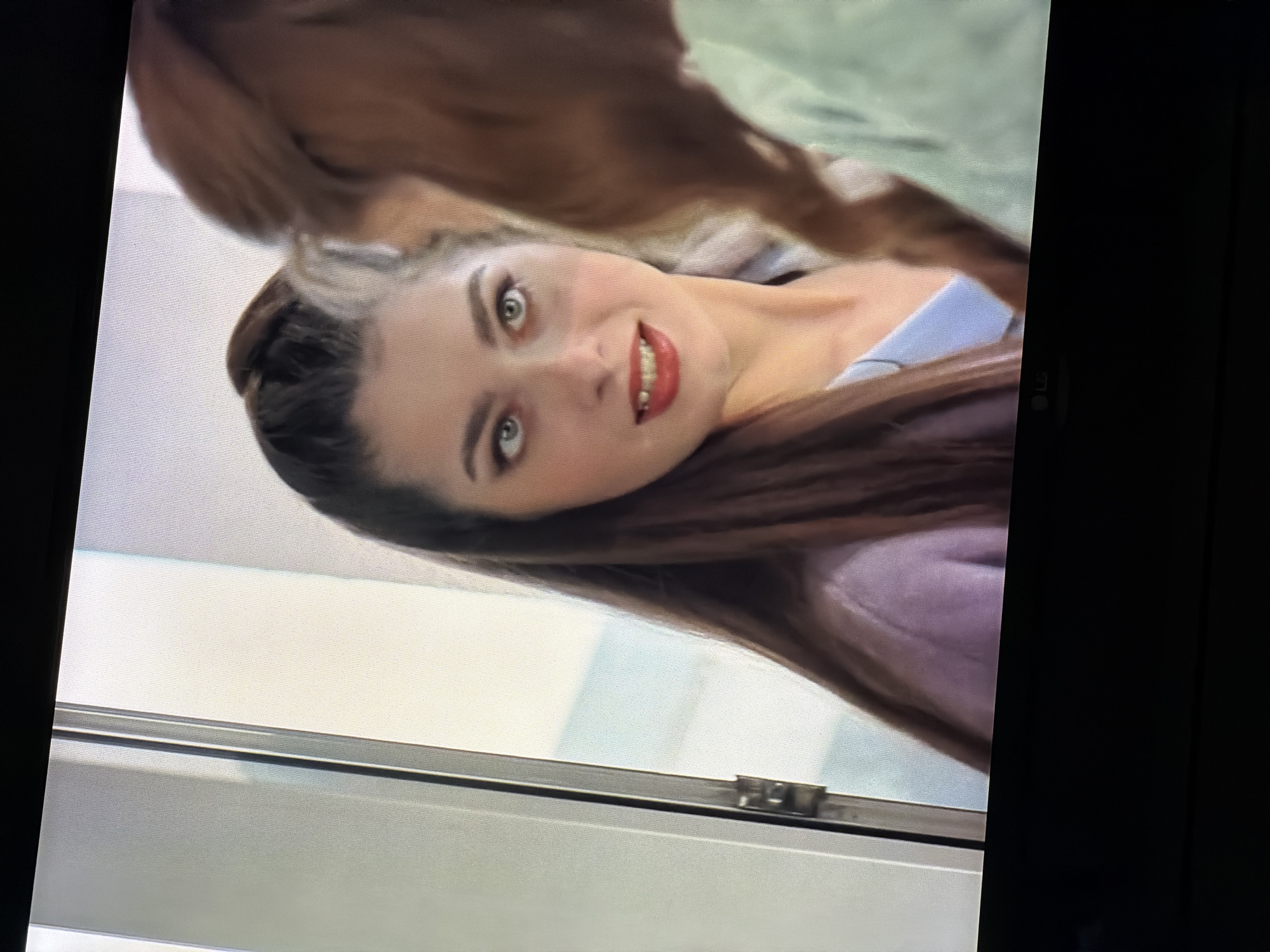 The image shows a medium close-up of two women indoors. The woman in the foreground has long, dark brown hair pulled into a high ponytail, with the rest of her hair falling around her shoulders. She is looking towards the right with a slight smile and her mouth is open, revealing her teeth. She is wearing makeup, including bright red lipstick and eyeliner. She is wearing a light purple sweater over a blue collared shirt.
The woman in the background is partially visible on the right side of the frame, her head and upper body are blurred. She has light brown hair and is wearing a light green garment.
The background consists of a bright, neutral-colored wall and what appears to be a door frame or window frame on the left side of the image. The lighting is bright and natural, suggesting an indoor setting during the daytime.
There is no visible indication or information within the image that suggests this scene is located in the city of Semiluki, Russia.