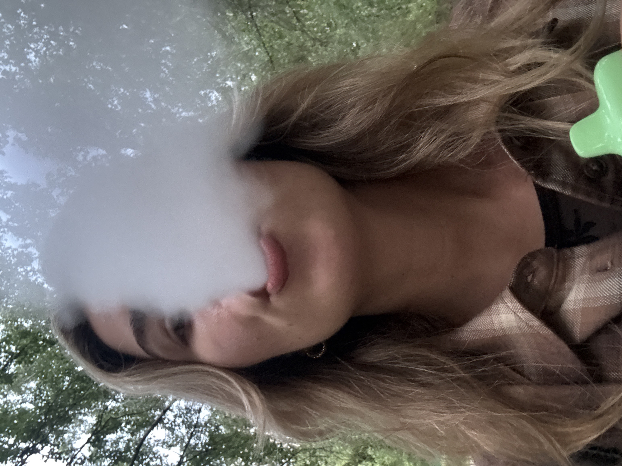 A young woman with blonde hair exhales a cloud of white vapor. She is wearing a plaid shirt and a black t-shirt underneath. Her lips are pursed, and she is looking to her left. A bright green object, possibly a vape pen, is visible in the lower right corner of the image. The background consists of green foliage and trees, suggesting an outdoor setting, possibly a park or wooded area. The lighting is natural, with dappled sunlight filtering through the leaves. The overall mood is casual and relaxed.