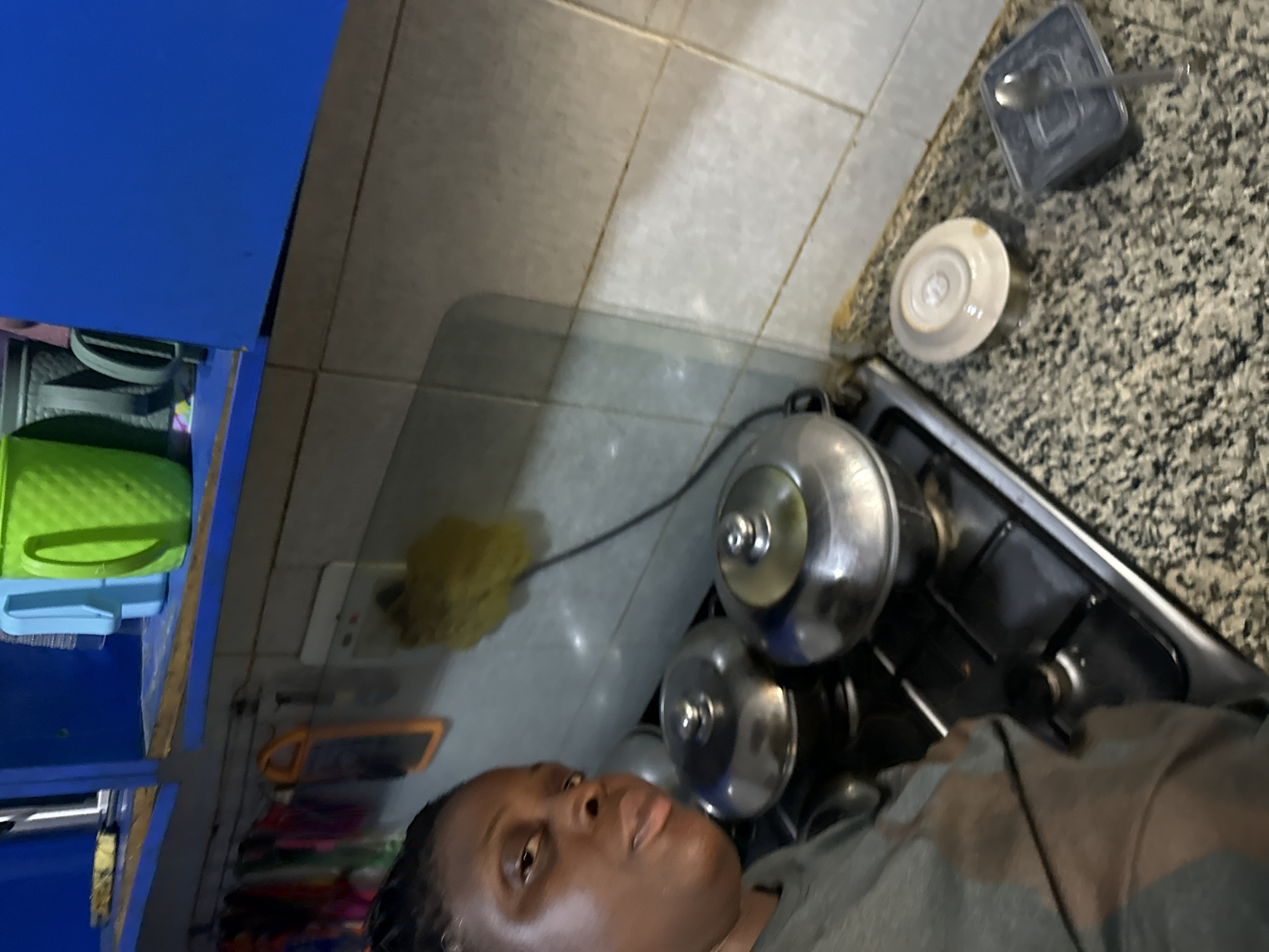 A photograph taken at eye level, in an interior location possibly in Sabon Tasha, Nigeria, depicts a woman standing in a kitchen. The kitchen includes blue cabinetry, white wall tiles and a counter top made of granite. The subject appears to be standing near a stove, on which sit metal pots and possibly a kettle. A white saucer and a covered rectangular container containing silverware are on the counter. A sponge and electrical outlet are nearby.