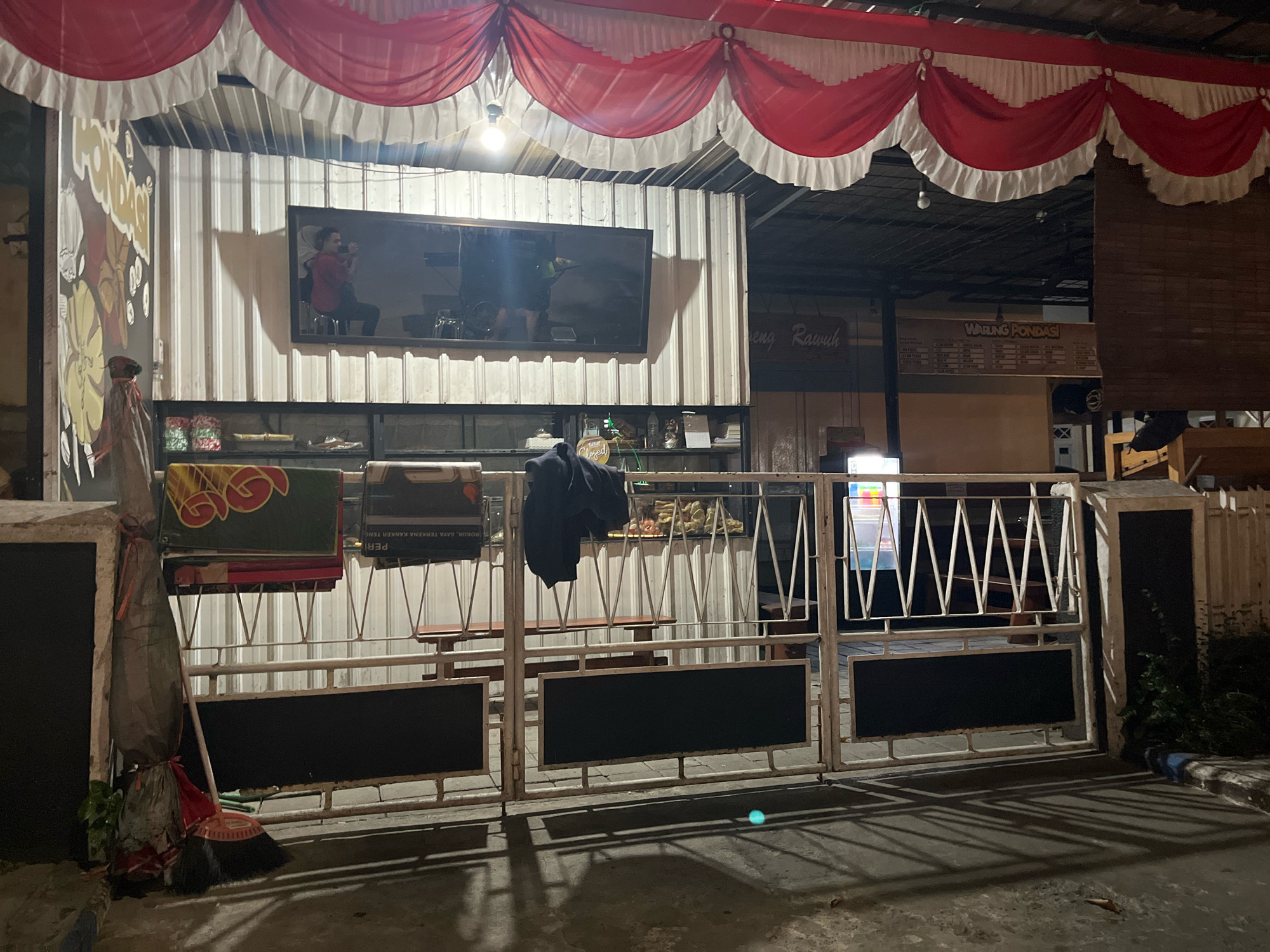 The image shows a storefront in Lumajang, Indonesia, at night. A large television screen is mounted on the front of the building, displaying an image of a person. Red and white curtains are draped across the top of the storefront. In front of the building, a white metal gate with black panels blocks access to the interior. A broom and a wrapped object are leaning against a post to the left of the gate. Various signs are visible on the building, including one that says "WARUNG PONDAS." The ground in front of the storefront is paved, and shadows are cast by the gate and other objects.