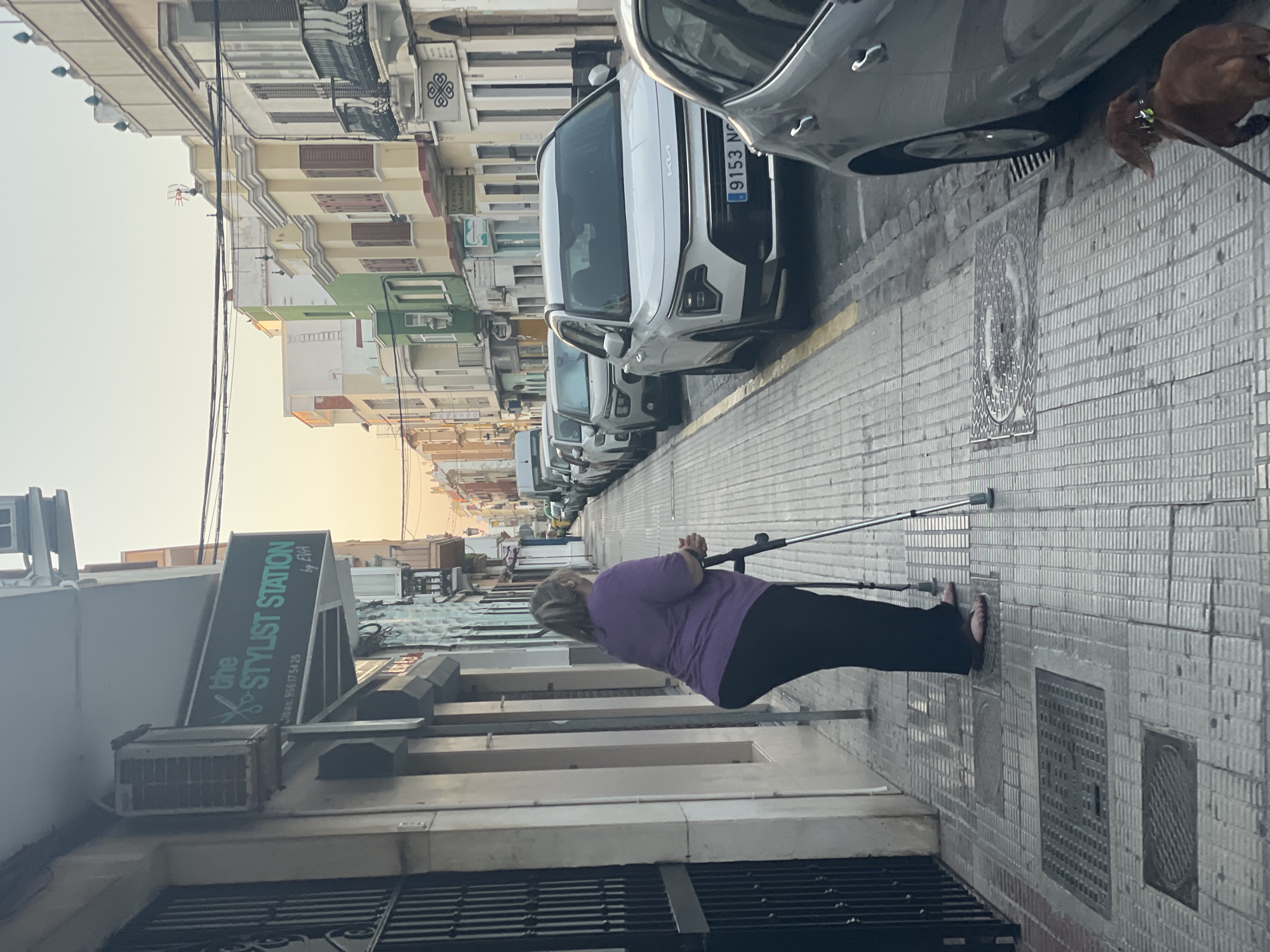 The media file depicts a street scene in La Línea De La Concepción, Spain. The main subject is a woman using a crutch for support as she walks on a tiled sidewalk. She is wearing a purple shirt and black pants. In the background, several cars are parked along the street, including a white Kia. Buildings with multiple stories line the street, displaying a variety of architectural styles and colors. A sign for "the STYLIST STATION by EVA" is visible on one of the buildings, along with a phone number: 956 17 54 25. The time of day appears to be late afternoon or early evening, with soft, warm lighting suggesting the golden hour. The weather seems clear. A brown dog on a leash can be seen in the upper right portion of the image. The scene conveys a sense of everyday life in a Spanish town.