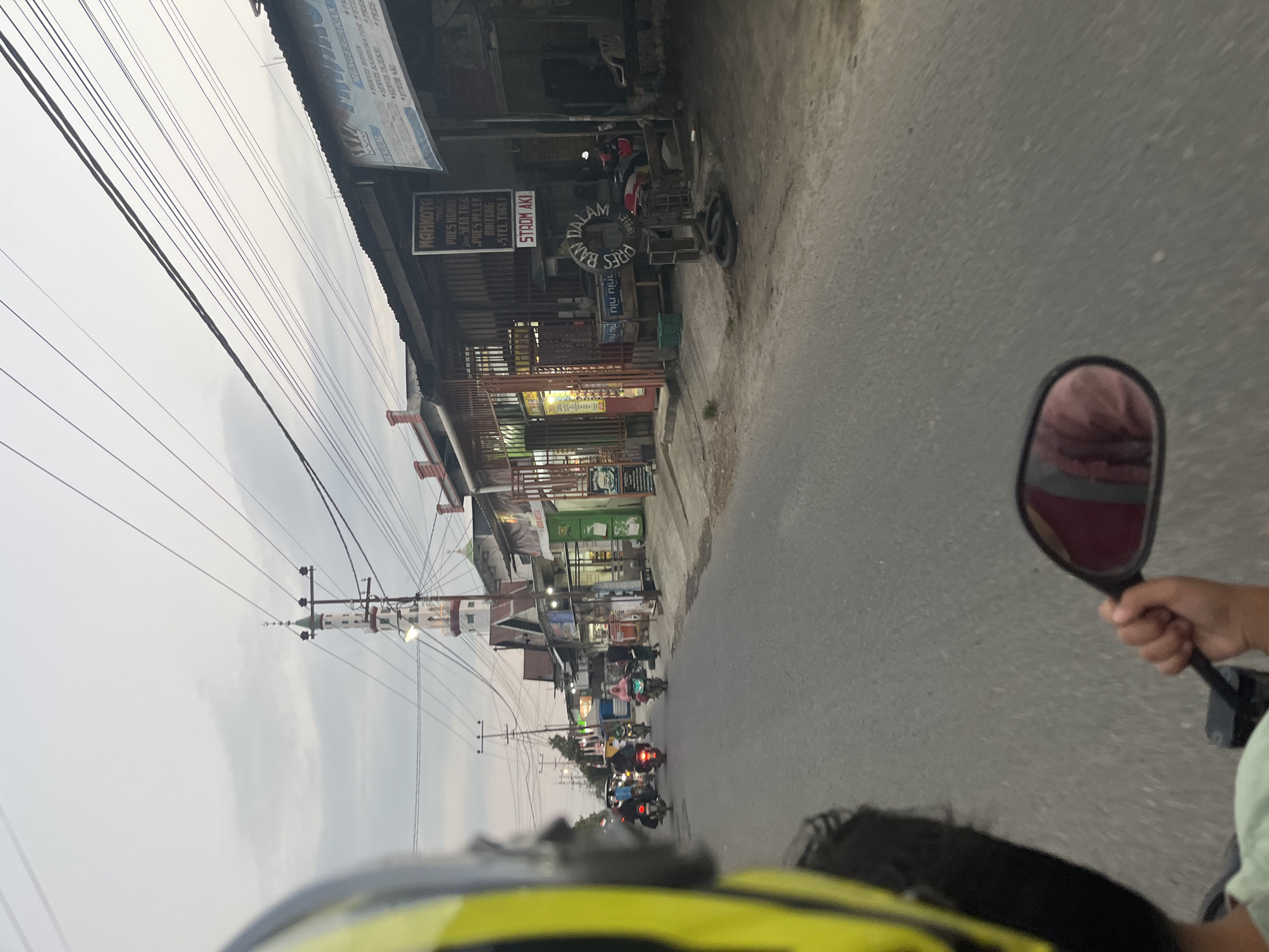 The image captures a view from the perspective of a motorcycle rider, looking down a paved road in Bumi Mulyo, Indonesia. The rider's yellow and black helmet is partially visible in the foreground, along with their hand holding a rearview mirror which reflects a person wearing a maroon garment.
The road is flanked by buildings on the right side, which appear to be shops or businesses. One building has a sign that reads "MAHKOTA" and lists services. Another sign displays "STROM AKI". Further down the road, a mosque with a tall, slender minaret stands prominently against the overcast sky. Several other motorcycles with riders, some wearing hijabs, are seen traveling in the same direction. Electrical wires crisscross above the road and buildings. The overall scene depicts a typical street in a semi-urban or rural area during daytime, with evidence of local commerce and religious infrastructure.