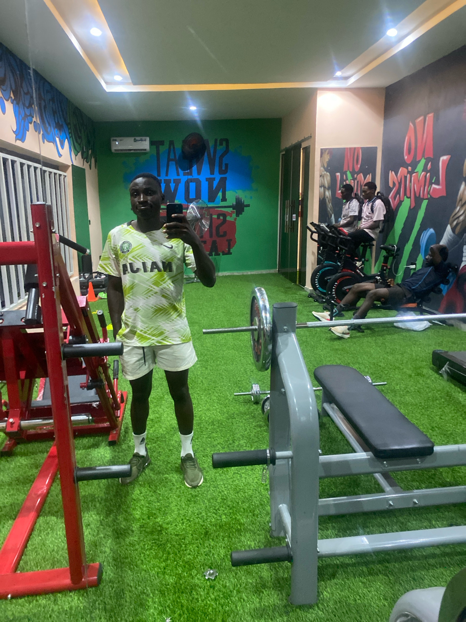 The image depicts the interior of a gym, likely located in Barden Yana, Nigeria. A young man is taking a mirror selfie in the foreground, wearing a white and green patterned t-shirt with "NIGERIA" emblazoned on it and white shorts. He is standing in front of a red weight machine. In the background, two other men are exercising on stationary bikes. The gym features green turf flooring, painted murals on the walls with motivational phrases like "SWEAT NOW" and "NO LIMITS", and various weightlifting equipment, including a bench press with weights. The lighting appears to be artificial, with ceiling lights illuminating the space.
