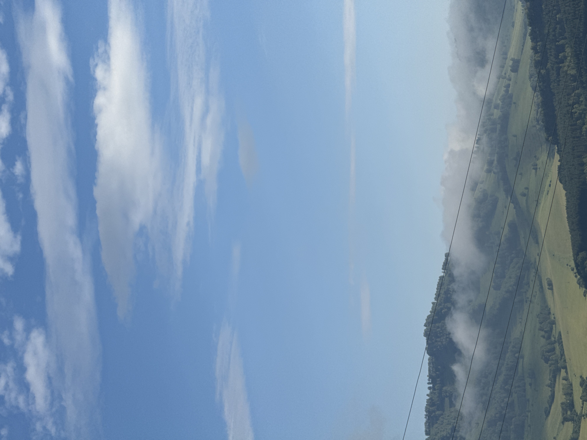 A scenic view of rolling green hills partially obscured by low-lying clouds and mist. Several power lines stretch horizontally across the frame, cutting through the landscape. The sky is a clear blue with scattered white clouds. The image appears to be taken from an elevated position looking out over a valley. The location is the city of Soloneshnoye, Russia.