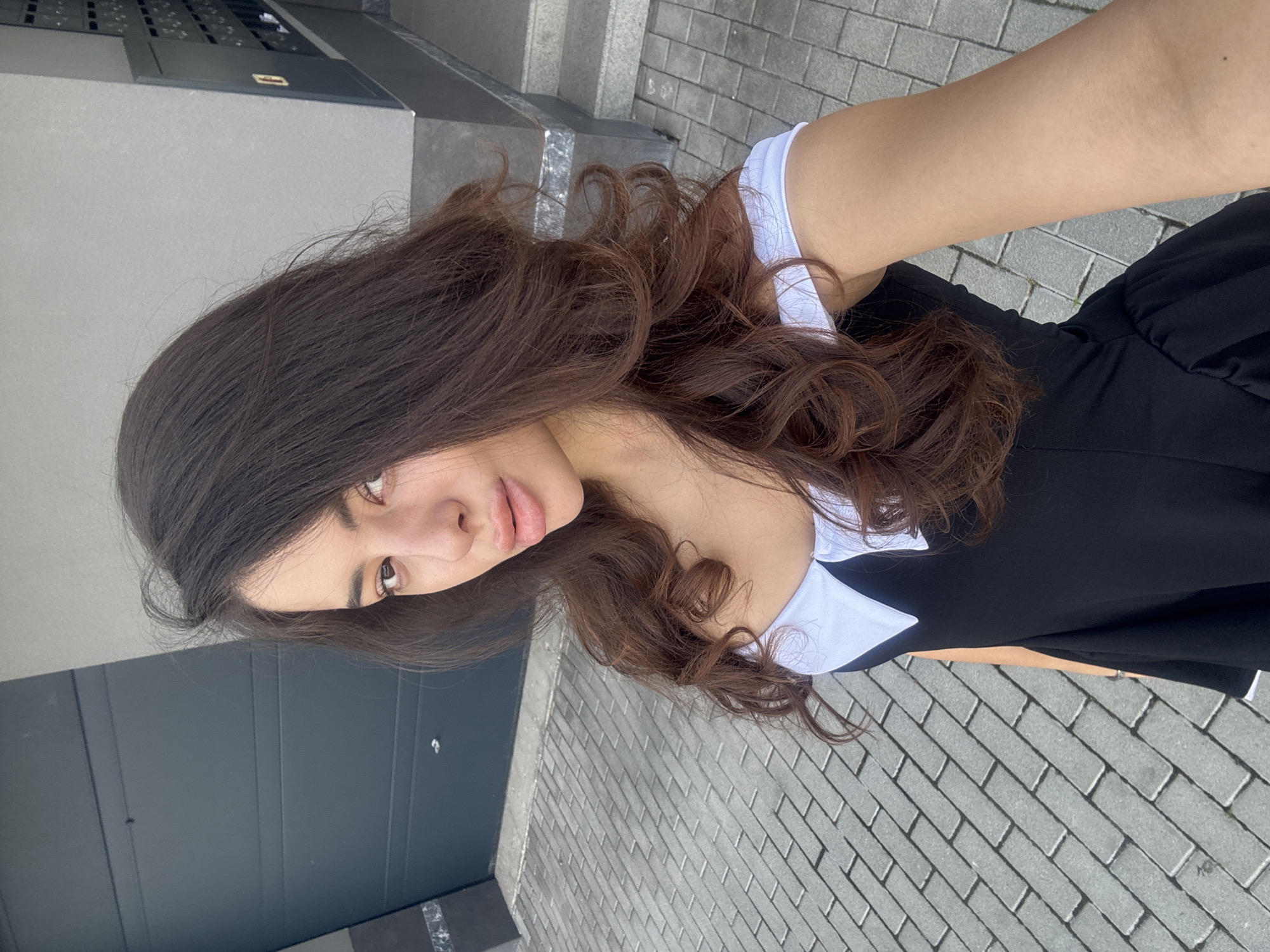 A young woman with long, dark, wavy hair and light brown eyes is taking a selfie outdoors. She is wearing a black dress with a white collar and short sleeves. The background shows a grey, textured wall with a dark gray garage door on the left and a set of stairs with dark gray metal railings on the right. In front of the building is a grey brick paved walkway. The woman is standing on the walkway, with the building behind her. The lighting suggests it is daytime.